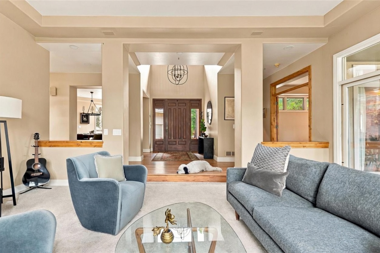 This inviting living room features a comfortable seating area with a blue sofa and matching armchair arranged around a glass-topped coffee table. The room opens into a grand, high-ceilinged entryway with a dark wood front door and a dog resting on the hardwood floor. The neutral color palette and open-concept layout create a spacious and welcoming atmosphere, with glimpses into adjacent rooms.