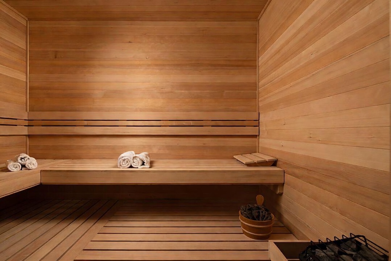 This image showcases a traditional sauna interior constructed with light-toned wood paneling on the walls and benches. The space includes a built-in bench, a wooden bucket with stones, and rolled towels, creating a serene and spa-like atmosphere. The perspective is a straight-on, eye-level view that emphasizes the clean lines and warm, natural aesthetic of the room.