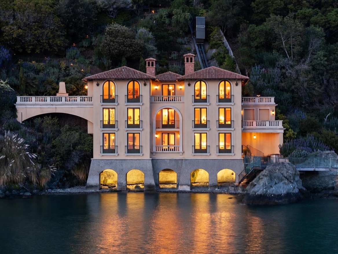 This striking Mediterranean-style villa is positioned directly on the water's edge, featuring a symmetrical facade with arched windows and multiple balconies. The structure is built into a steep, lush hillside, connected to the upper terrain by a private funicular railway. The warm glow from the interior lights reflects beautifully on the calm water, creating a serene and luxurious atmosphere.