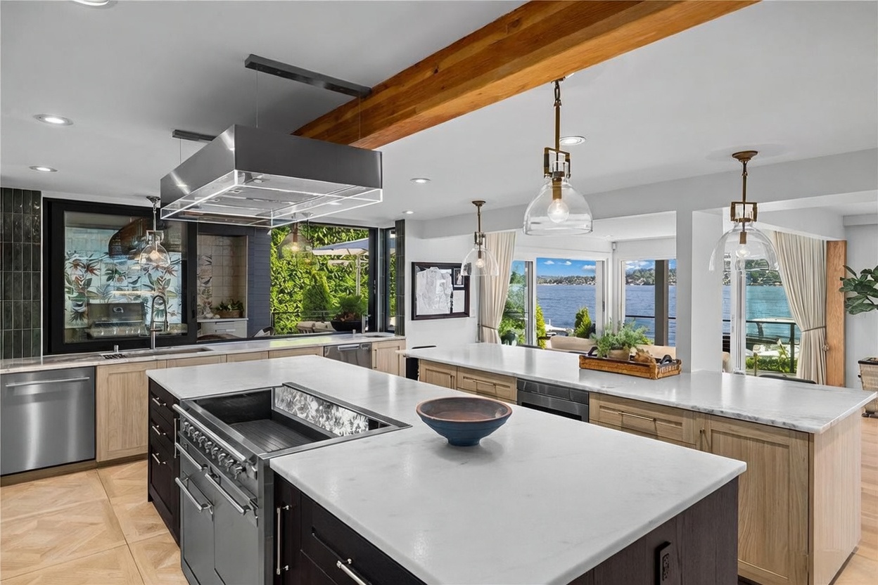 This expansive, modern kitchen features two large islands with light-toned stone countertops and dark cabinetry, creating a sophisticated contrast. A prominent wooden beam spans the ceiling, while multiple pendant lights hang above the islands, illuminating the workspace. Large glass doors open to a scenic waterfront view, seamlessly blending the indoor cooking area with the outdoor environment.