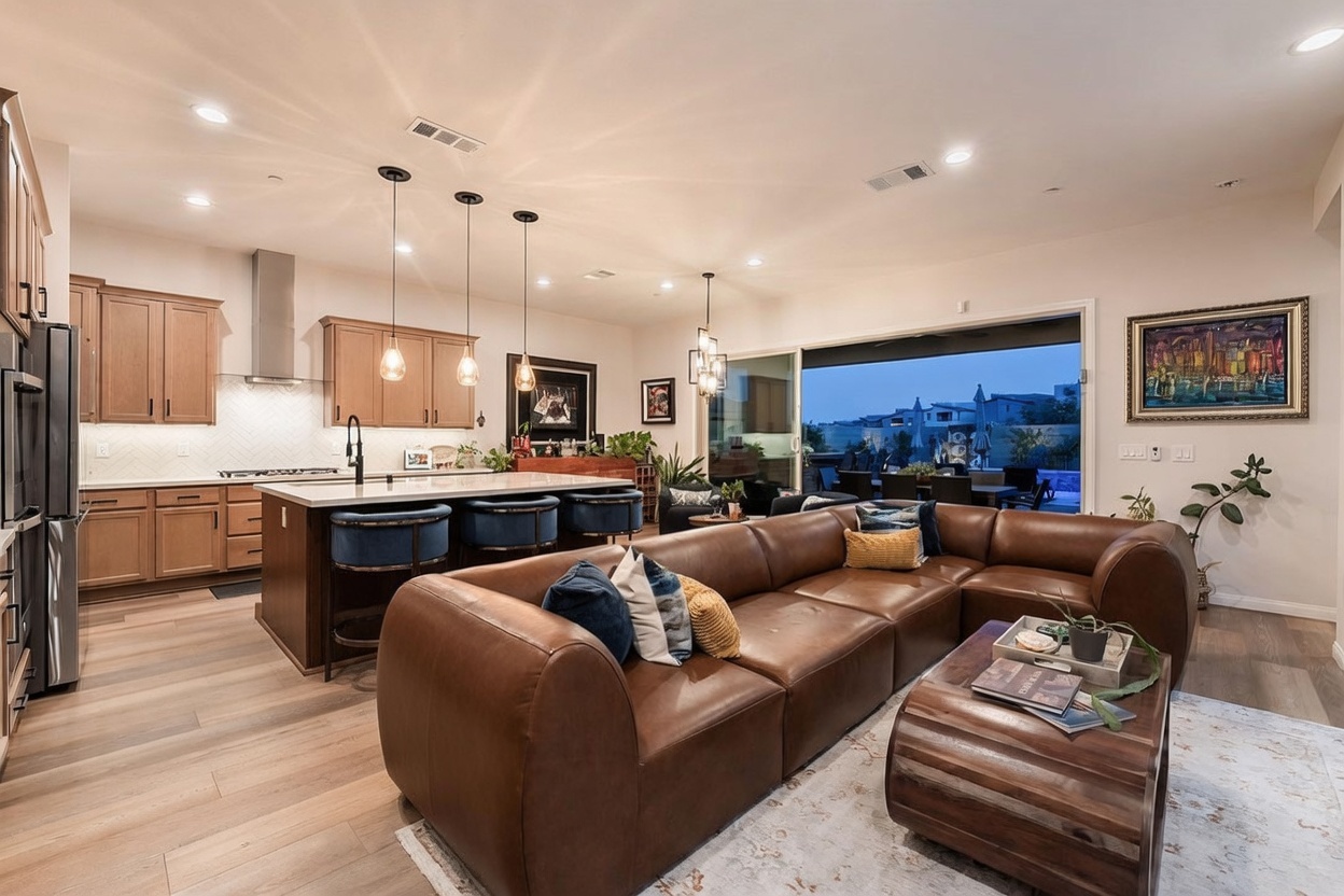 This open-concept living space features a large, plush brown leather sectional sofa arranged around a rustic wooden coffee table. The room flows seamlessly into a modern kitchen equipped with light wood cabinetry, a central island with bar seating, and pendant lighting. Large sliding glass doors open to an outdoor patio, creating a bright and airy atmosphere that blends indoor and outdoor living.