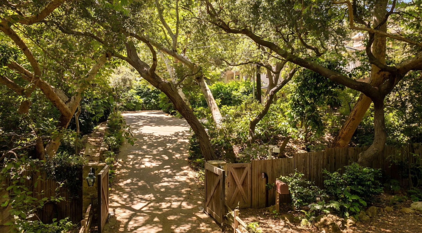 This image captures a private, gated driveway entrance framed by mature, sprawling trees that create a canopy of dappled light. A rustic wooden gate stands open, leading to a paved path that winds toward a secluded residence nestled in the lush greenery. The setting offers a serene, wooded atmosphere, emphasizing privacy and a natural, park-like approach to the property.