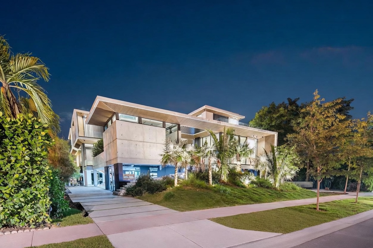 This striking modern residence features a bold architectural design with exposed concrete walls, clean lines, and expansive glass elements. The home is set on a landscaped lot with a paved driveway leading to a covered entrance, illuminated by warm exterior lighting against a twilight sky. The combination of natural textures and contemporary geometry creates a sophisticated and inviting curb appeal.