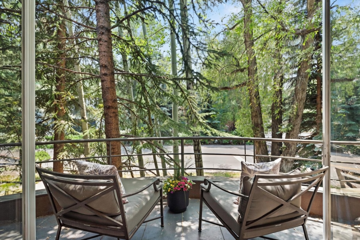 This balcony offers a serene, elevated view of a lush, wooded area through a metal railing. Two comfortable outdoor chairs with patterned cushions are positioned on either side of a vibrant potted plant, creating an inviting space for relaxation. The perspective is from inside looking out through large glass panels, emphasizing the seamless connection between the indoor living space and the natural surroundings.