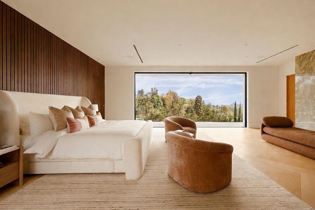 This spacious primary bedroom features a large, light-colored upholstered bed set against a textured wood-paneled wall. A expansive window provides a clear view of the surrounding trees, while two curved armchairs and a low-profile bench create a comfortable seating area on the light wood flooring. The room is designed with a minimalist, modern aesthetic that emphasizes natural light and clean lines.