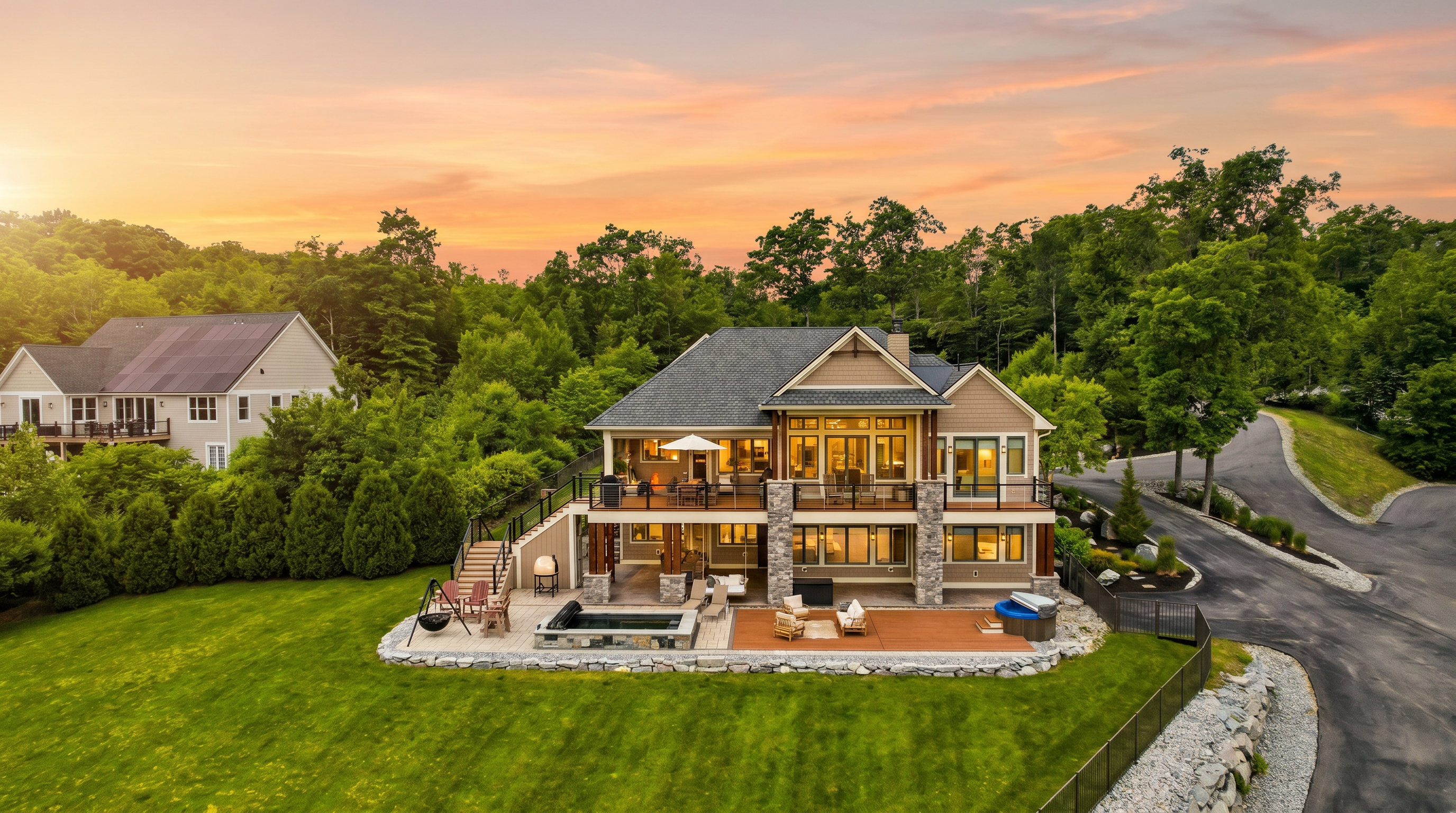 This elevated rear view captures a multi-level luxury home set against a lush, wooded backdrop. The property features a spacious upper deck and a lower-level patio complete with a built-in pool, hot tub, and outdoor seating area, all overlooking a manicured lawn. The architecture blends stone and siding elements, creating a sophisticated retreat that seamlessly integrates indoor and outdoor living spaces.