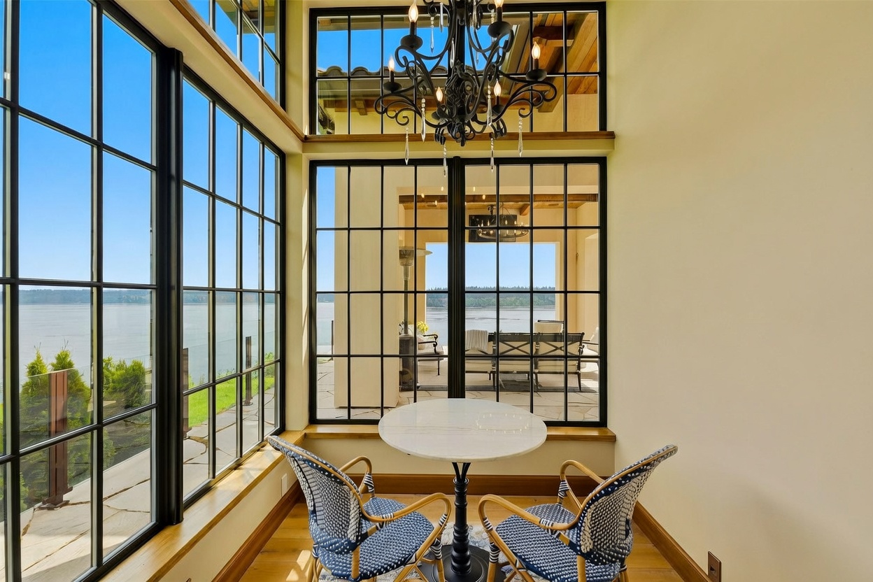 This elegant sunroom features expansive, black-framed windows that offer panoramic views of the water and an adjacent outdoor patio. The space is furnished with a small, round stone-topped table and two woven chairs, all centered beneath a decorative black chandelier. The room is finished with warm-toned wood flooring, creating a bright and inviting atmosphere perfect for relaxation.