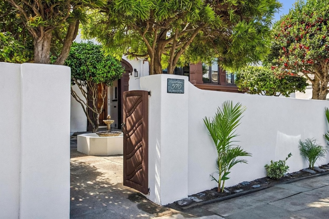 This inviting entryway features a crisp white stucco wall and a decorative wooden gate that opens into a private courtyard. A tiered stone fountain serves as a focal point near the entrance, surrounded by lush greenery and mature trees that provide dappled shade. The scene is captured from a street-level perspective, highlighting the home's charming curb appeal and gated privacy.