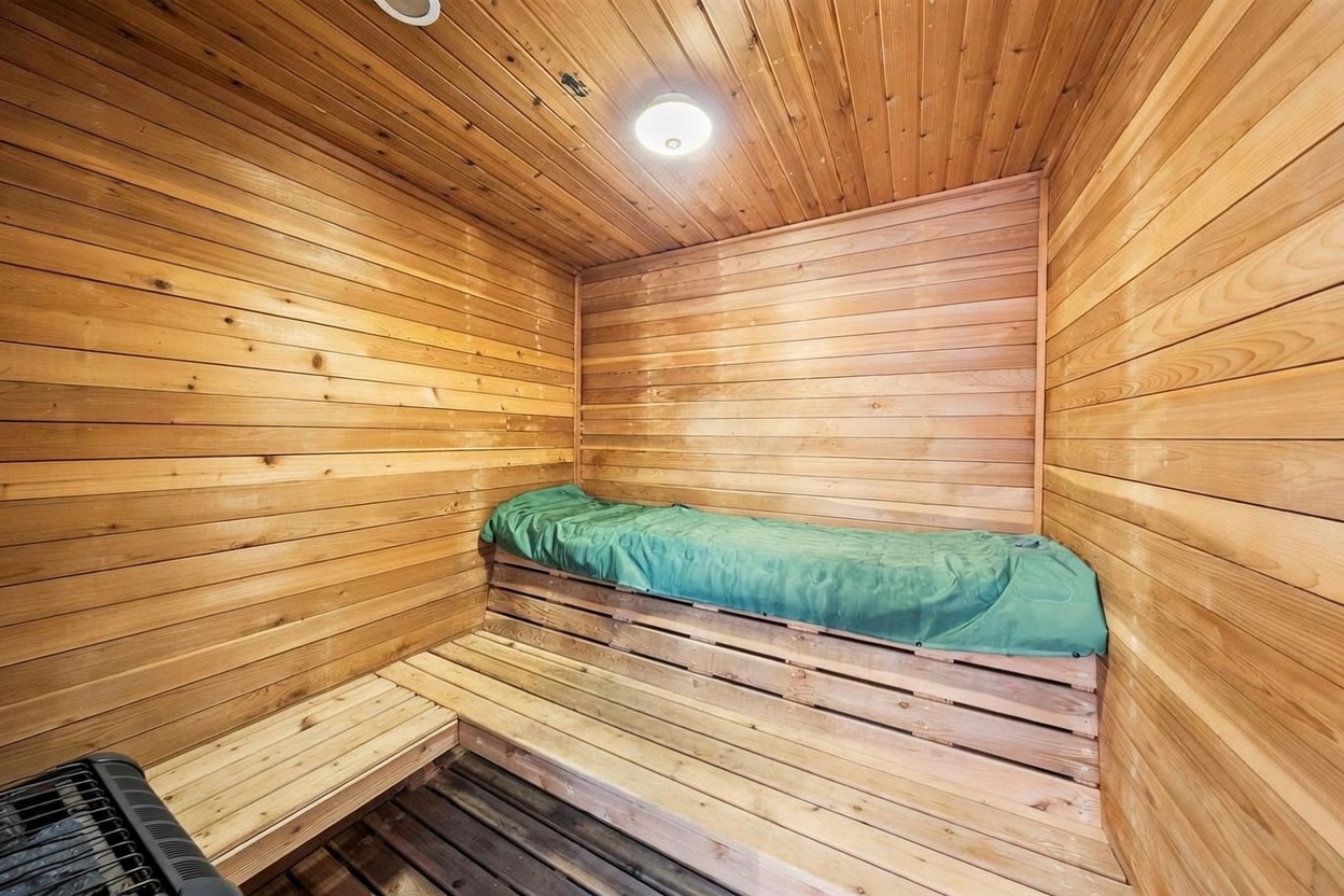 This image shows a compact, wood-paneled sauna room featuring horizontal cedar planks on the walls and ceiling. A built-in wooden bench with a green cover provides seating, while a metal heater unit is visible in the foreground. The warm, natural wood tones create a cozy and relaxing atmosphere typical of a private home spa amenity.
