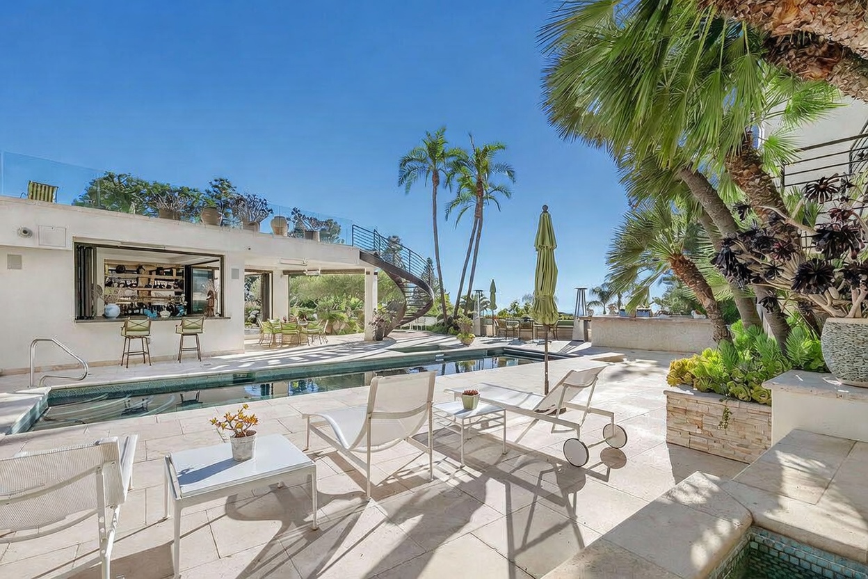 This luxurious outdoor living space features a long, rectangular swimming pool as the central focal point, surrounded by light-toned stone decking. To the left, an open-air bar area with stools provides a seamless transition from the interior, while a curved staircase leads to an upper terrace. Tall palm trees and manicured landscaping frame the scene, creating a serene and upscale resort-like atmosphere under a clear blue sky.