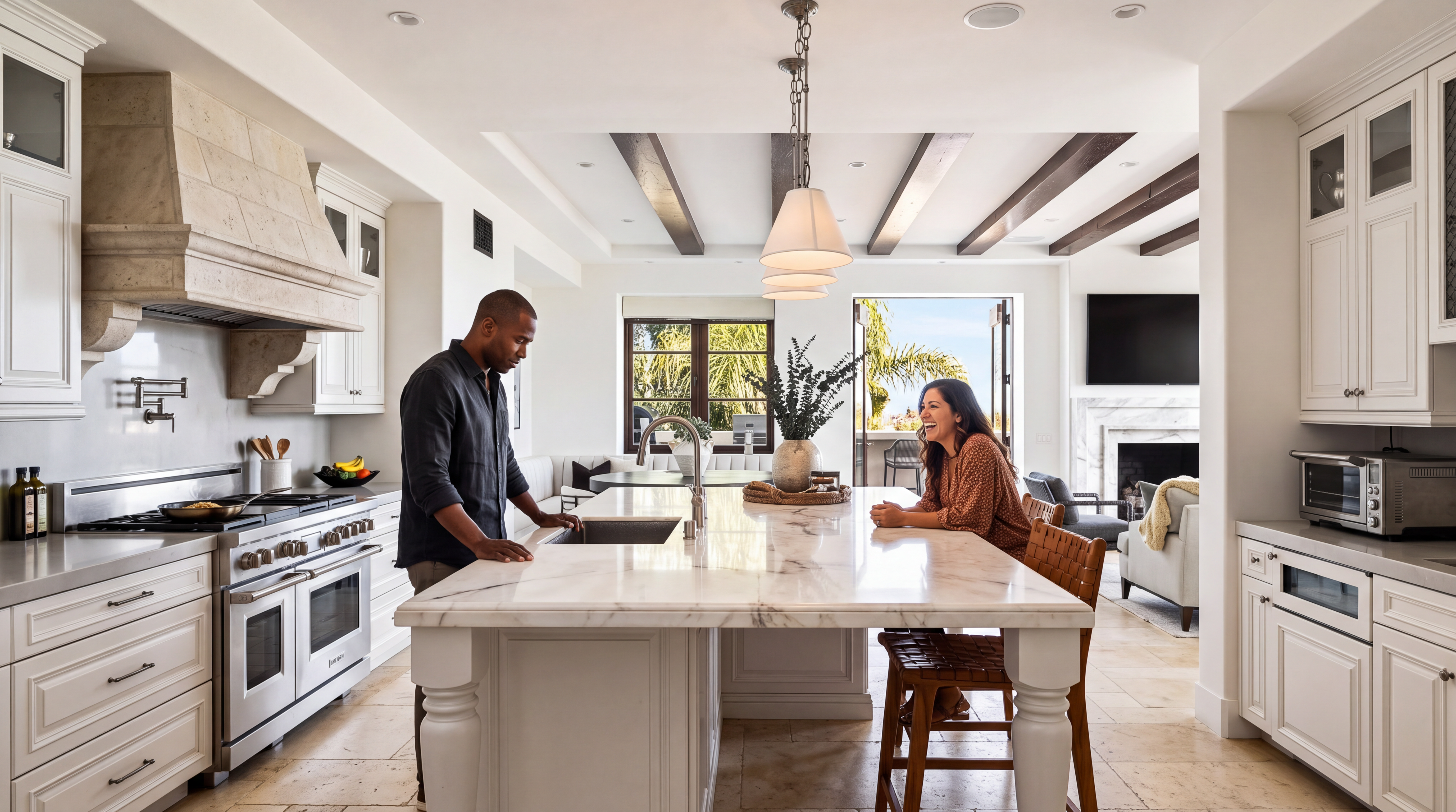 This spacious, light-filled kitchen features high-end marble countertops, an expansive central island with a farmhouse sink, and custom off-white cabinetry. Exposed wood ceiling beams add character and warmth to the room, which seamlessly flows into an open-concept living area with a cozy fireplace. The space is elegantly styled with modern lighting and high-quality appliances, creating a welcoming and sophisticated environment for entertaining.