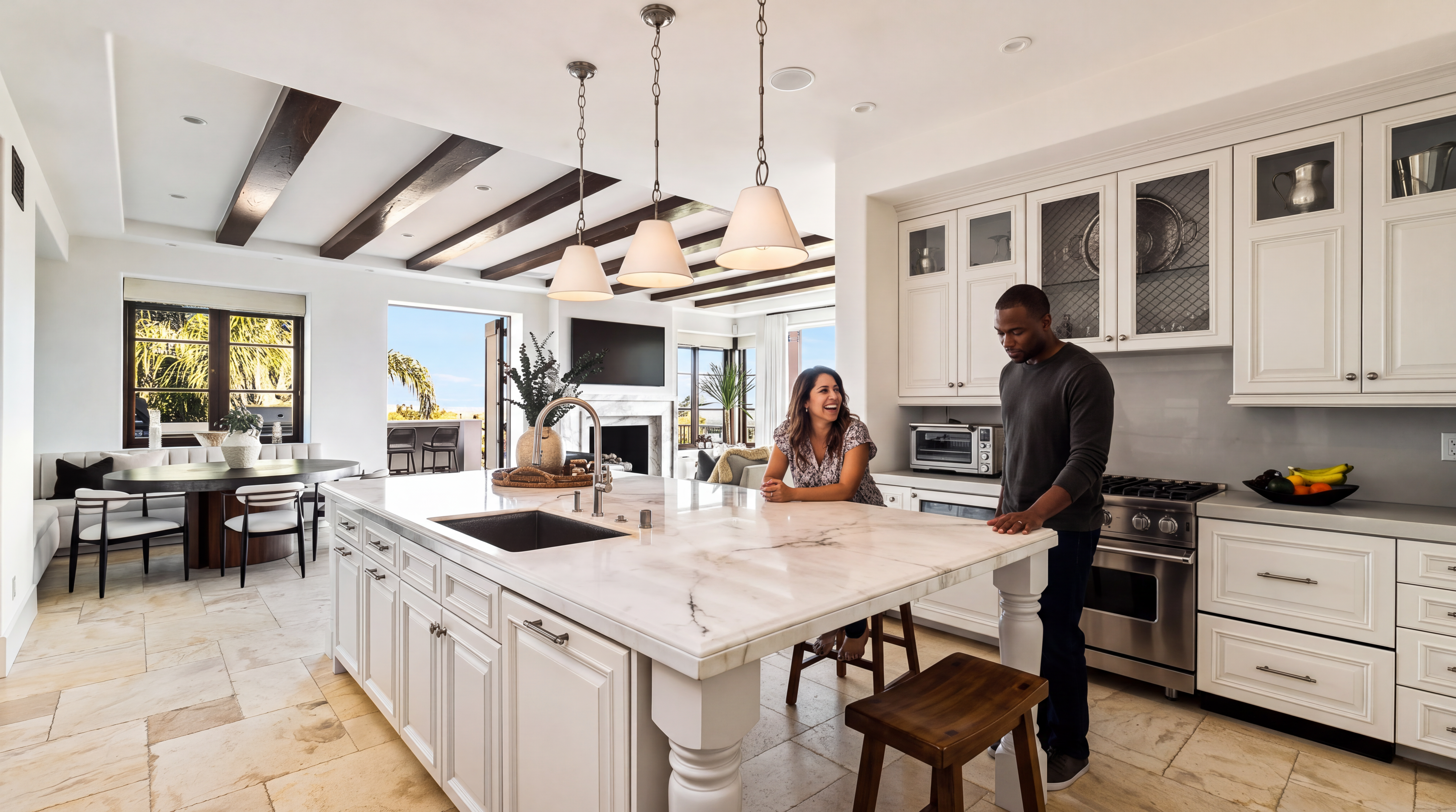This bright, open-concept kitchen features a large marble-topped island and classic white cabinetry, creating a sophisticated and welcoming space. It boasts high-end stainless steel appliances, decorative pendant lighting, and exposed dark ceiling beams that add architectural character. With a seamless transition to the dining area and an outdoor view, this kitchen is designed for both elegant entertaining and comfortable daily living.