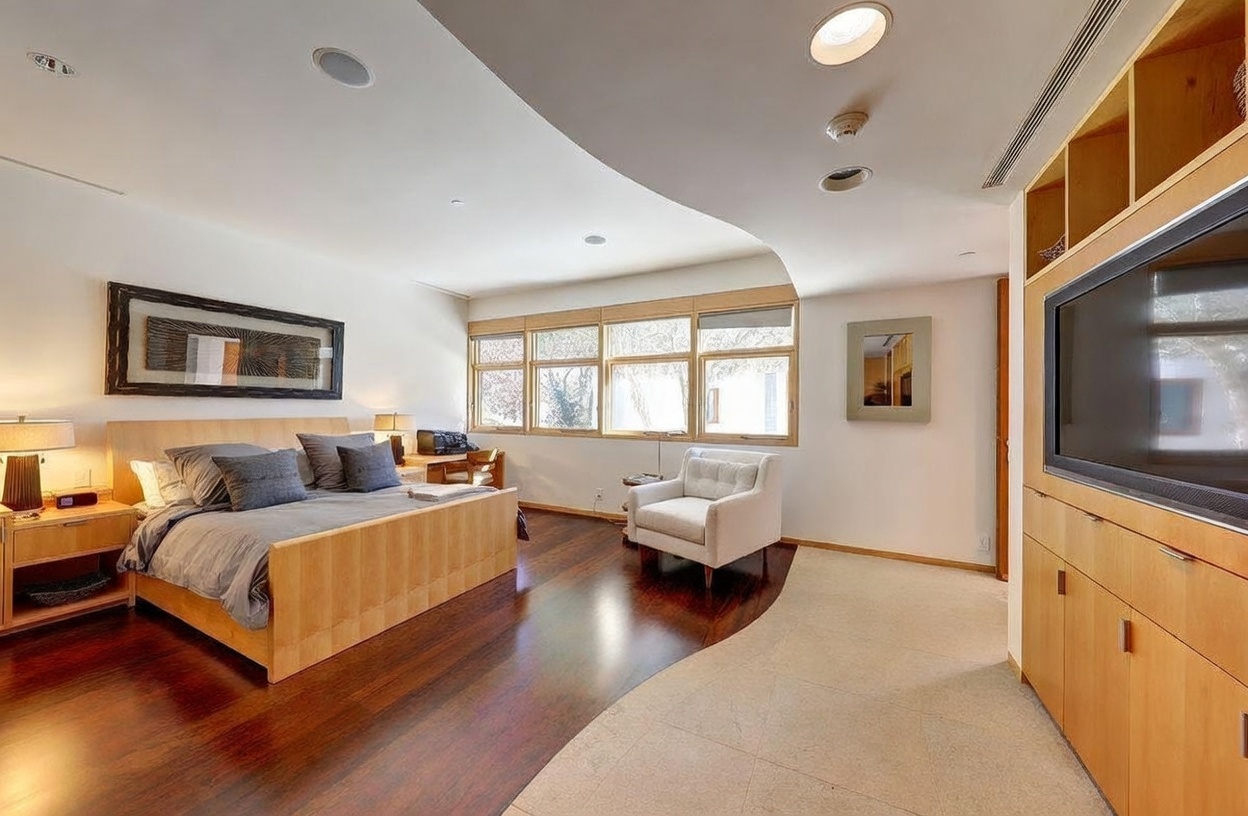 This spacious primary bedroom features a distinct transition between dark wood flooring in the sleeping area and light-colored tile in the adjacent sitting area. The room is furnished with a light wood bed frame, a white armchair, and a built-in media cabinet, all illuminated by recessed lighting and natural light from a series of windows. The curved ceiling design adds a unique architectural element to the room's contemporary aesthetic.