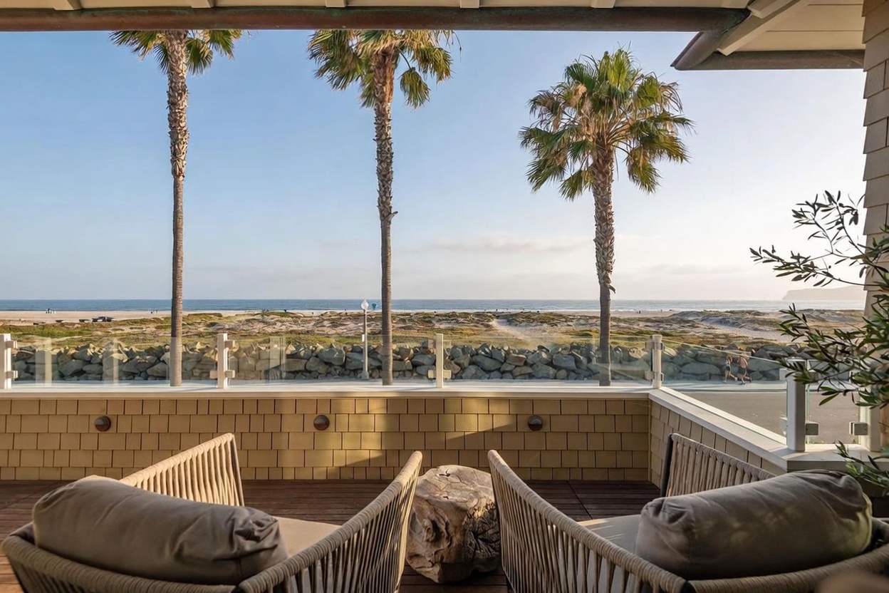 This elevated balcony offers a serene, direct view of the beach and ocean, framed by three tall palm trees. The space is furnished with two comfortable, woven-back lounge chairs and a rustic, natural wood stump side table, creating a relaxed coastal atmosphere. A glass railing ensures an unobstructed sightline to the sandy dunes and shoreline below, emphasizing the property's prime waterfront location.