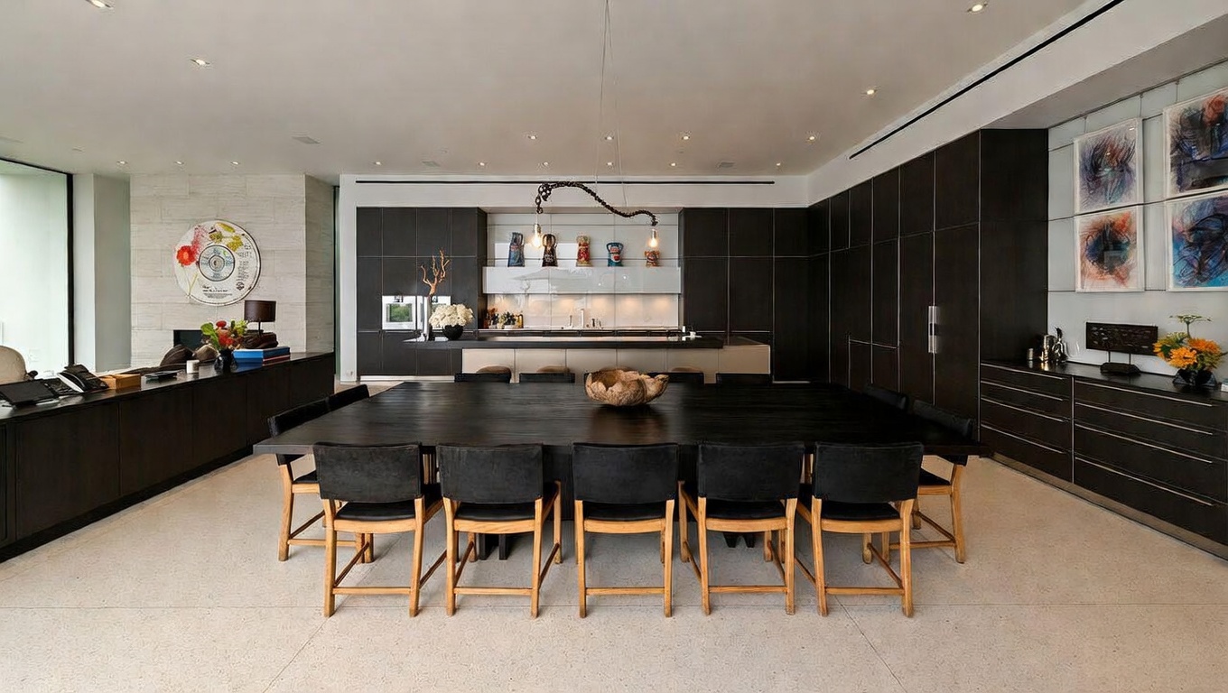 This sophisticated dining area features a large, dark wood table as its centerpiece, surrounded by minimalist chairs with light wood frames. The space seamlessly integrates with a sleek, modern kitchen in the background, characterized by dark cabinetry and a clean, open-concept layout. The room is accented by contemporary artwork and recessed lighting, creating an elegant and spacious atmosphere perfect for formal entertaining.