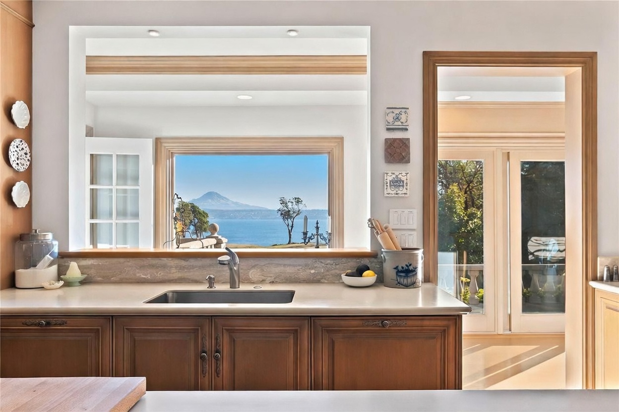 This kitchen features warm-toned wood cabinetry and a neutral stone countertop with an integrated sink. A large, open window frame provides a stunning, unobstructed view of a lake and a distant mountain peak, serving as a natural focal point. To the right, a doorway leads to an adjacent room, while decorative wall tiles and a ceramic utensil crock add a touch of character to the space.