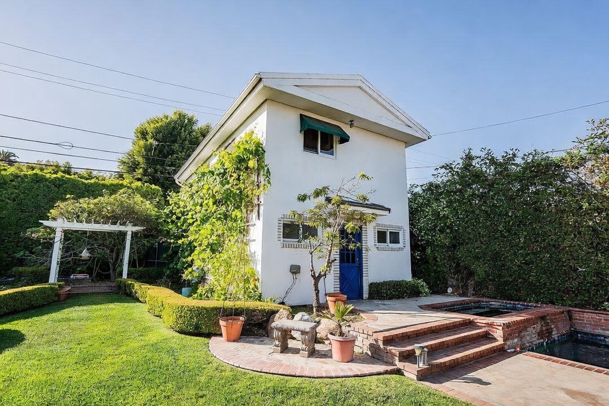 This exterior view captures a charming two-story guest house or accessory dwelling unit set within a lush, private backyard. The structure features white siding, a blue door, and a small green awning, complemented by a brick-edged pool and spa area in the foreground. A manicured lawn, neatly trimmed hedges, and a white wooden pergola create a serene, park-like atmosphere perfect for outdoor relaxation.