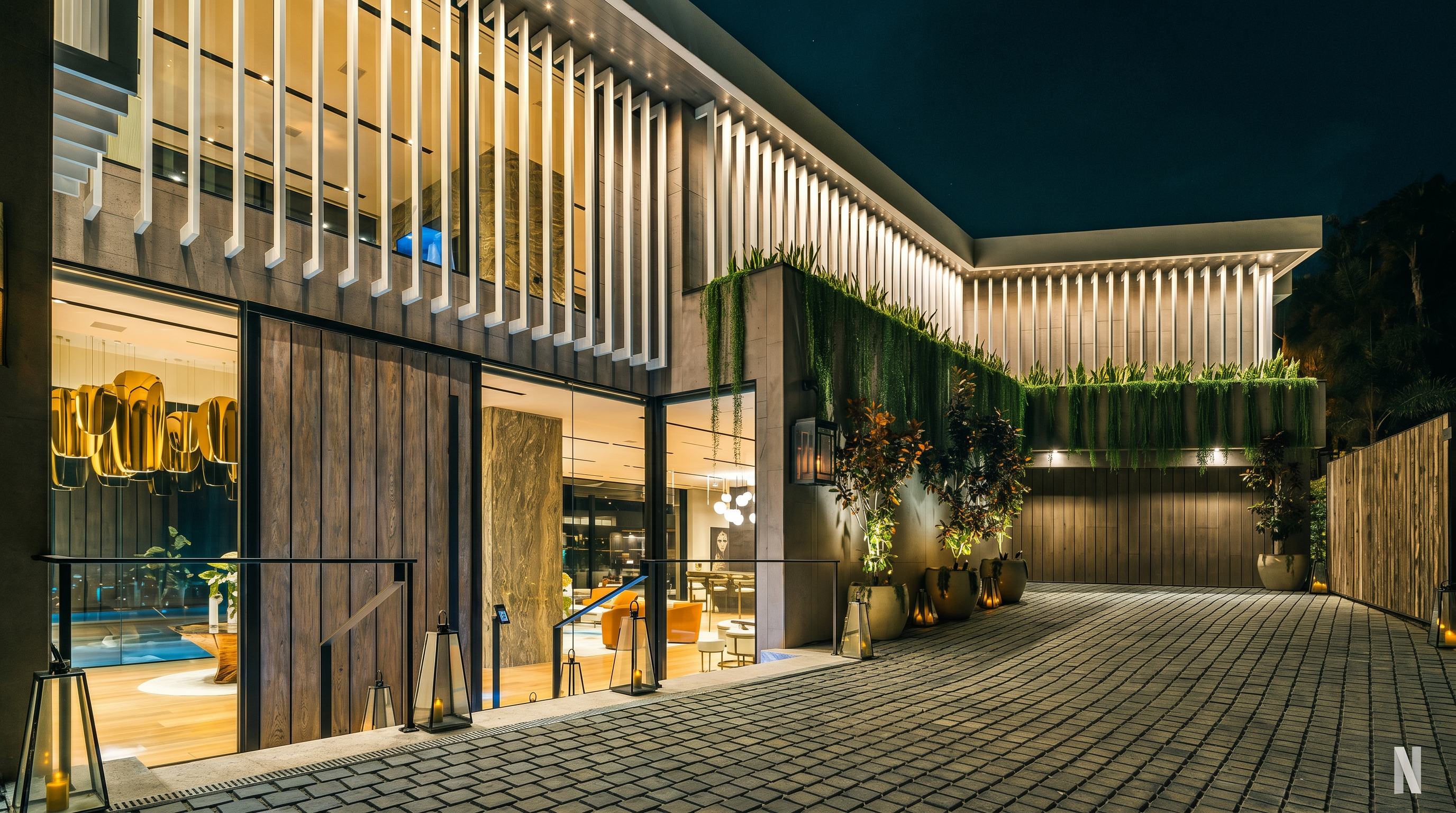 This striking modern entryway features a sophisticated blend of vertical wood paneling, floor-to-ceiling glass, and dramatic architectural fins that create a rhythmic facade. The paved driveway leads to a warm, inviting entrance illuminated by elegant lanterns and recessed lighting, while lush greenery cascades from the upper level. The overall aesthetic is one of high-end luxury, emphasizing clean lines and a seamless transition between indoor and outdoor living spaces.
