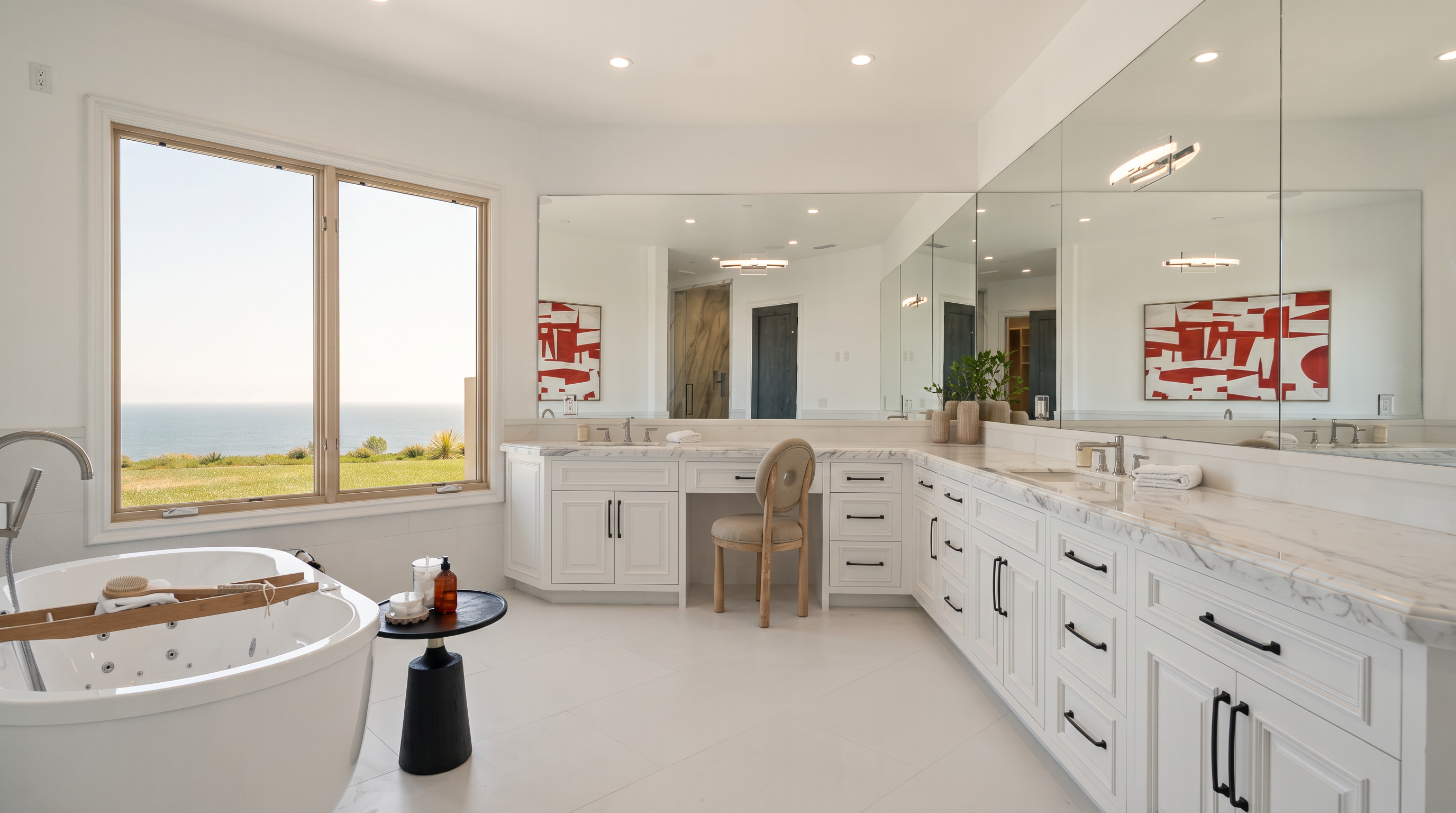 This spacious primary bathroom features a large soaking tub positioned near expansive windows that offer a panoramic ocean view. The room is anchored by a long, white vanity with stone countertops and a built-in makeup desk, complemented by a large wall-to-wall mirror. The bright, neutral color palette and clean lines create a serene and luxurious atmosphere.