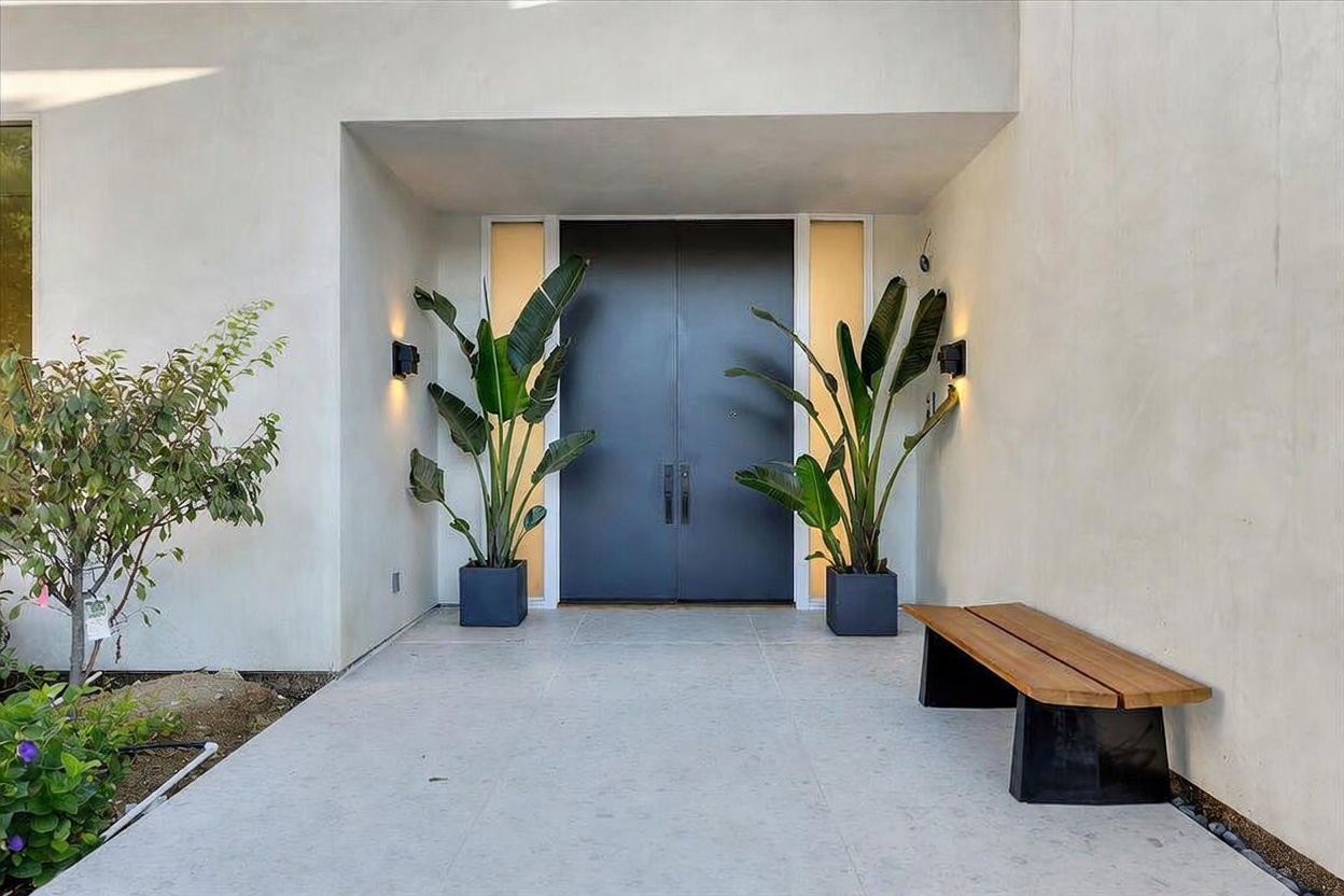 This modern entryway features a pair of dark, sleek double doors flanked by tall, narrow frosted glass panels. Two large potted plants sit symmetrically on either side of the entrance, while a minimalist wooden bench rests on the light-colored paved patio. The clean, smooth stucco walls and warm wall-mounted lighting create a sophisticated and welcoming atmosphere.