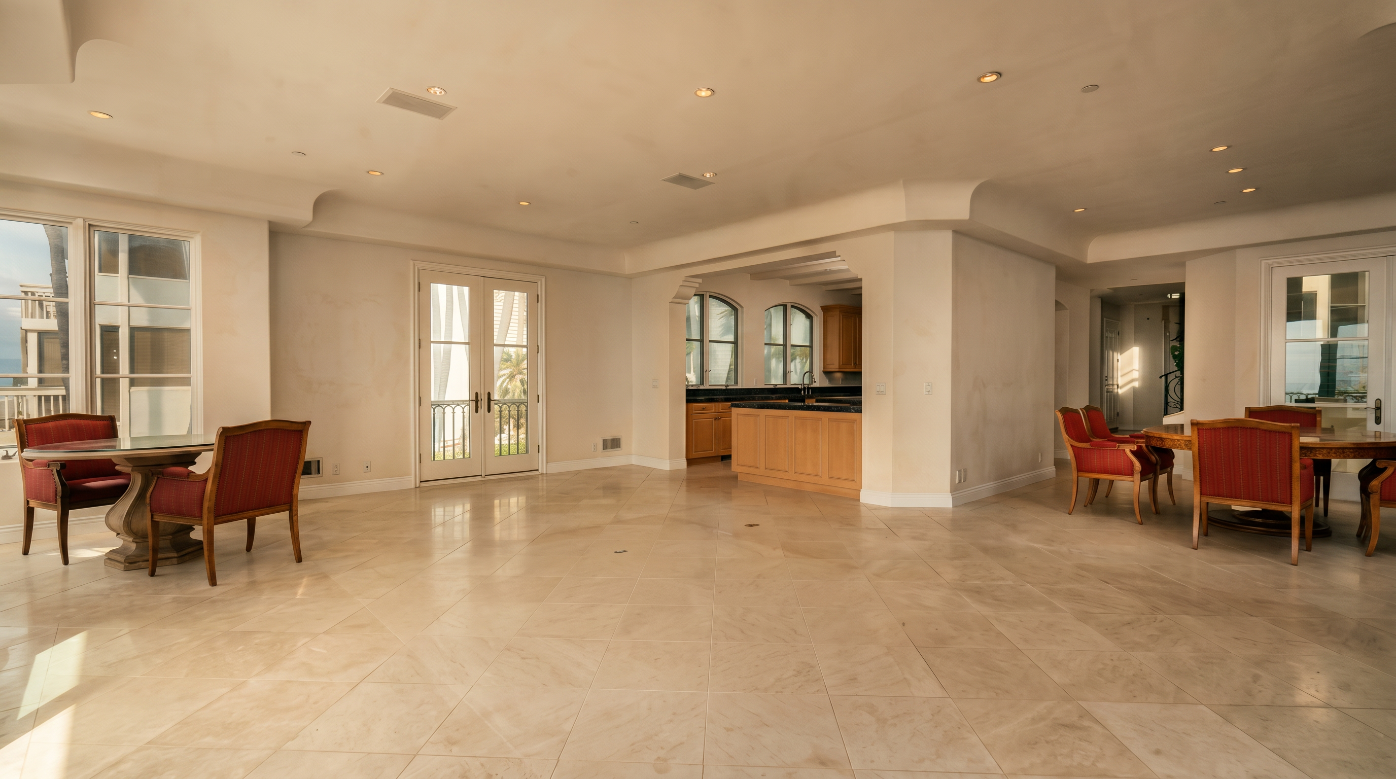 This spacious, open-concept living and dining area features light-colored tile flooring and neutral white walls, creating a bright and airy atmosphere. The room includes two distinct seating areas with red-upholstered chairs and round tables, while a kitchen with light wood cabinetry and a central island is visible in the background. Large glass doors and windows provide ample natural light and views of the outdoors, contributing to a clean and expansive aesthetic.