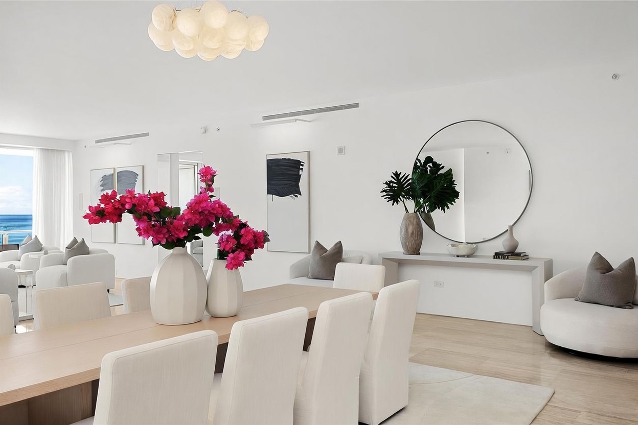 This bright and airy dining room features a long, light-toned table surrounded by modern, upholstered chairs. A striking floral arrangement with vibrant pink blossoms serves as a centerpiece, while a large circular mirror and minimalist artwork enhance the sophisticated, contemporary aesthetic. The space is illuminated by a unique, cloud-like pendant light fixture and benefits from natural light streaming in from the adjacent living area.