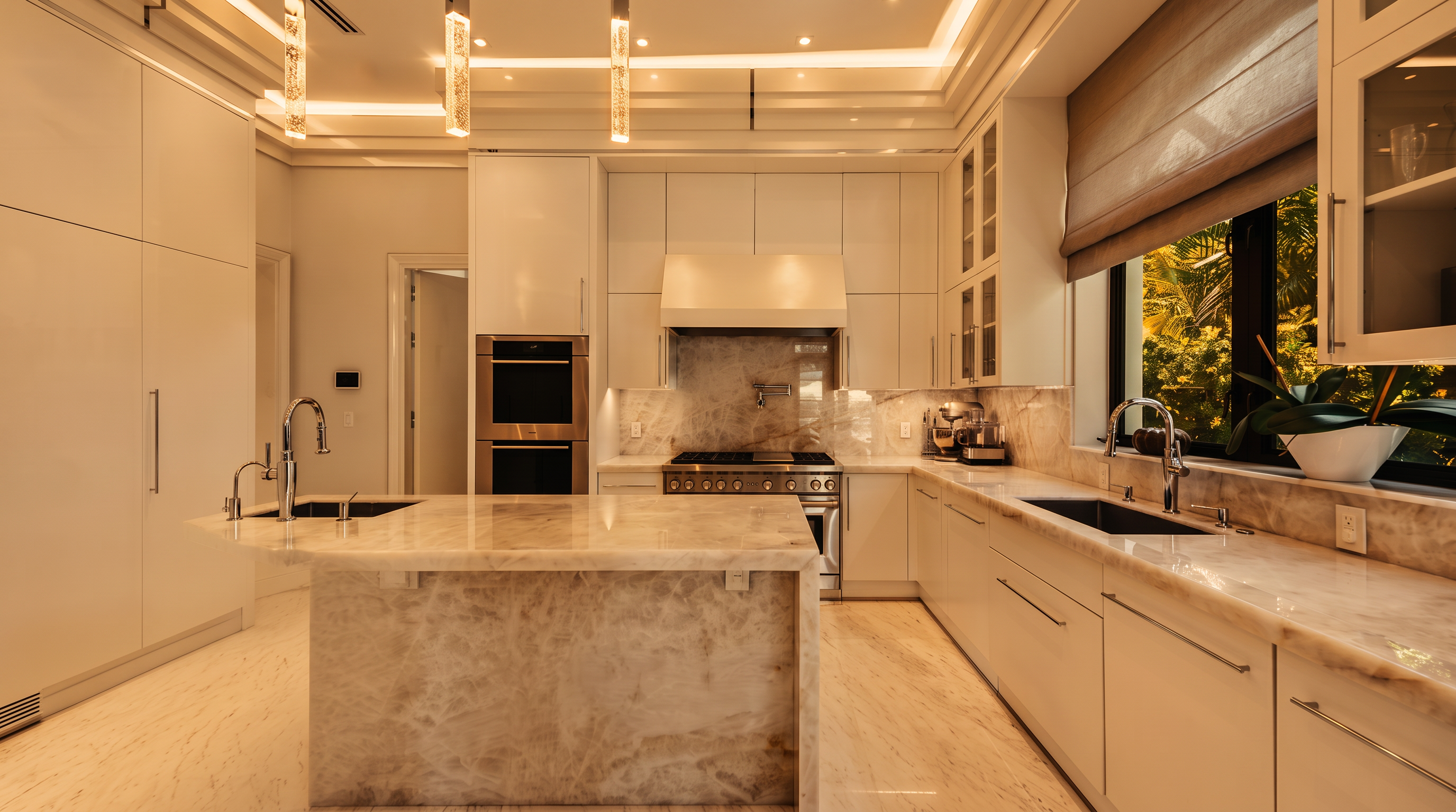 This bright, modern kitchen features sleek white cabinetry, a large central island with a stone waterfall edge, and high-end stainless steel appliances. The space is illuminated by minimalist pendant lights and recessed ceiling lighting, creating a clean and sophisticated atmosphere. A large window above the sink provides a view of lush greenery, while the neutral stone surfaces throughout add a sense of luxury and cohesion.
