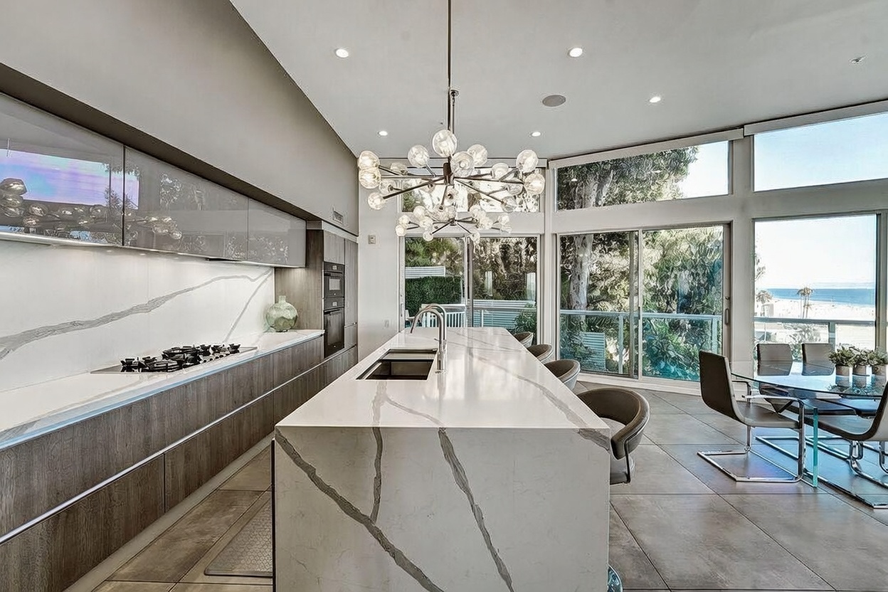 This modern, open-concept kitchen features a large central island with a waterfall-edge stone countertop and integrated sink, complemented by sleek, dark-toned cabinetry. A striking, multi-globe chandelier hangs above the island, while expansive glass sliding doors provide a seamless connection to the outdoors and offer a view of the beach. The space is finished with large-format floor tiles and minimalist design elements, creating a sophisticated and airy atmosphere.