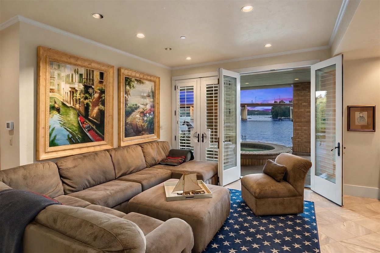 This inviting living room features a large, plush sectional sofa and a matching armchair arranged on a navy blue rug with a star pattern. The space is decorated with two framed landscape paintings and a decorative model sailboat on an ottoman, while glass doors open directly to a patio overlooking a waterfront view with a bridge in the distance. The room is illuminated by recessed lighting and offers a seamless transition between indoor comfort and the scenic outdoor environment.