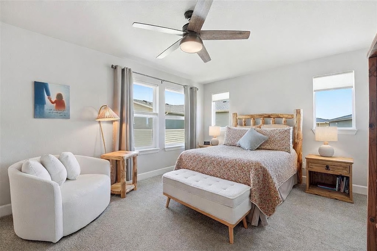 This cozy bedroom features a rustic log-style bed frame as the focal point, complemented by a patterned quilt and a tufted bench at the foot of the bed. To the left, a curved white armchair sits beside a small wooden side table and a floor lamp, creating a comfortable reading nook. The room is brightened by multiple windows with neutral curtains, light-colored walls, and a neutral carpet, offering a warm and inviting atmosphere.
