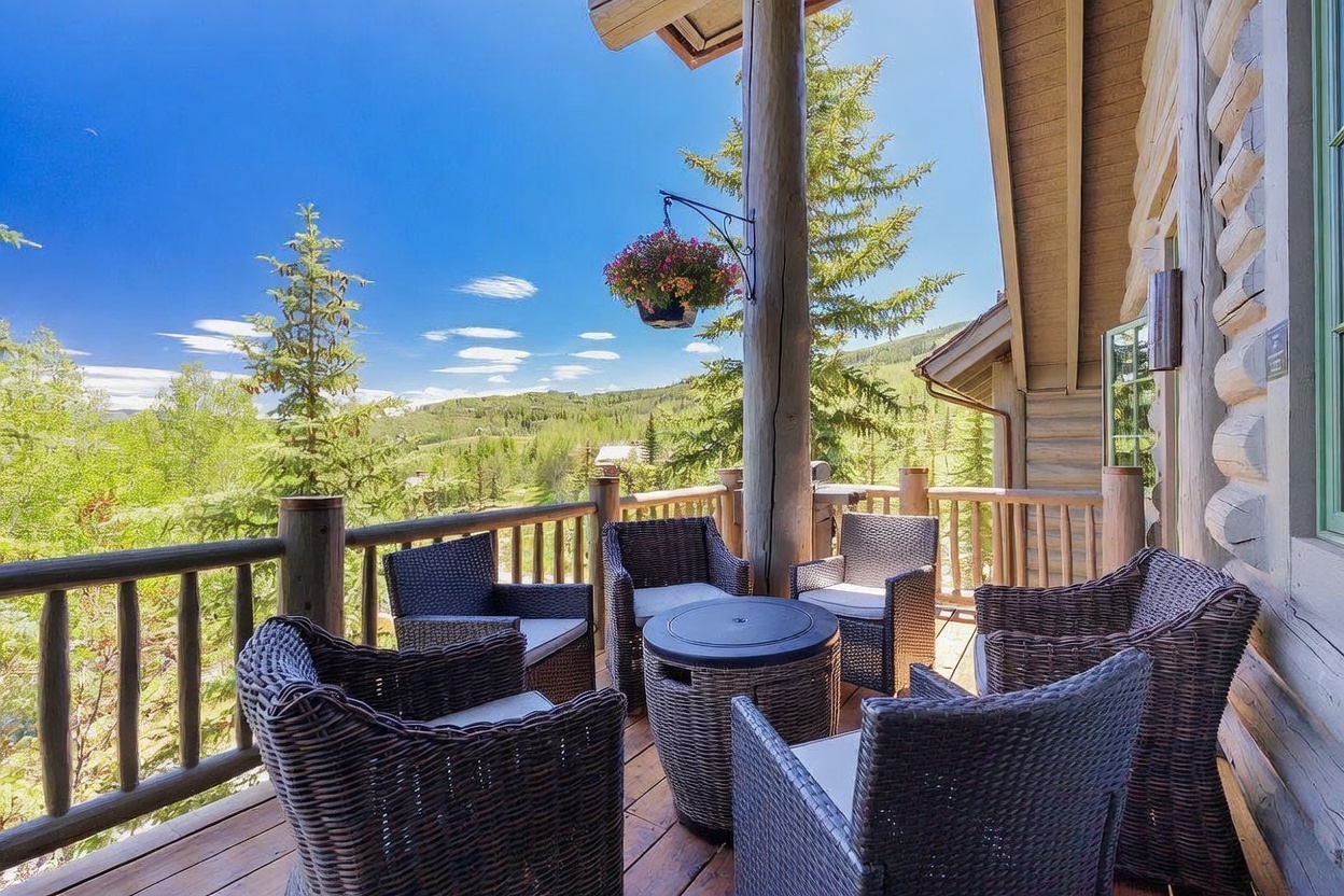 This elevated wooden deck offers a serene outdoor living space furnished with a circular fire pit table surrounded by four wicker-style armchairs. The deck features a sturdy wooden railing and is accented by a hanging flower basket, providing a cozy spot to enjoy the expansive mountain and forest views. The rustic log-style siding of the home adds to the mountain retreat aesthetic, creating a warm and inviting atmosphere.