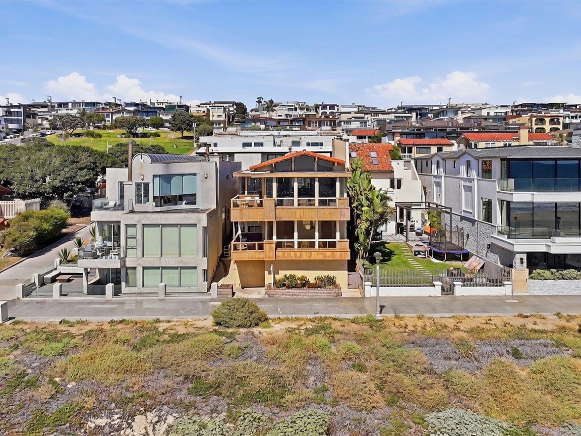 This aerial perspective captures a row of coastal homes situated along a paved walkway, with the central property featuring a distinctive wood-clad facade and multiple levels of balconies. To the right, a neighboring home includes a grassy yard with a trampoline, while the foreground is dominated by a natural, brush-covered coastal landscape. The scene conveys a relaxed, upscale beachside lifestyle with clear views of the surrounding neighborhood and oceanfront proximity.