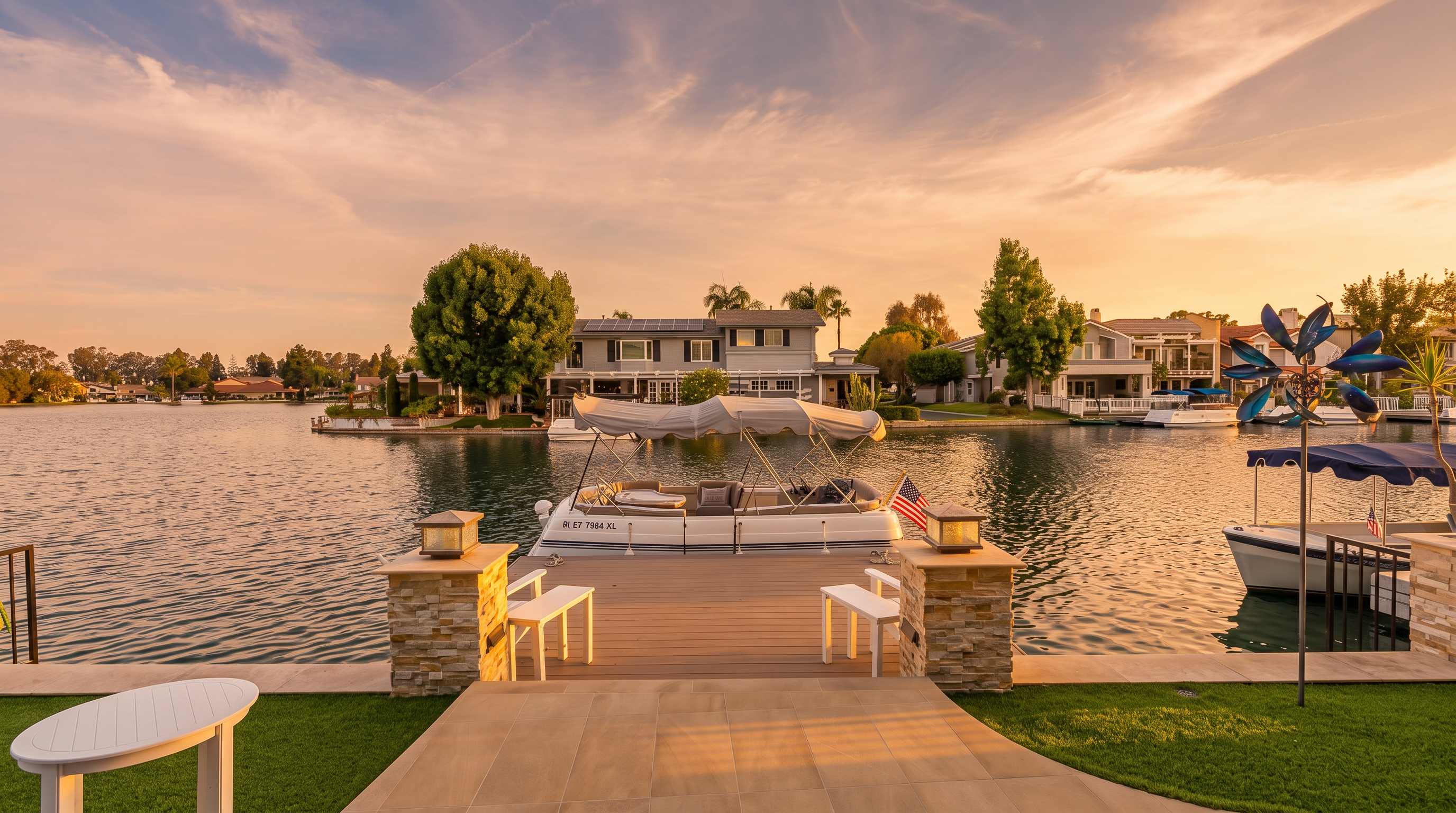 This stunning waterfront property features a pristine private dock with a docked pontoon boat on a calm, blue lake. The foreground includes a neatly tiled patio area with artificial turf, stone-capped pillars, and modern outdoor furniture, creating a luxurious and serene outdoor living space. The view showcases well-maintained houses along the shoreline, highlighting the attractive lifestyle of this lakeside home.