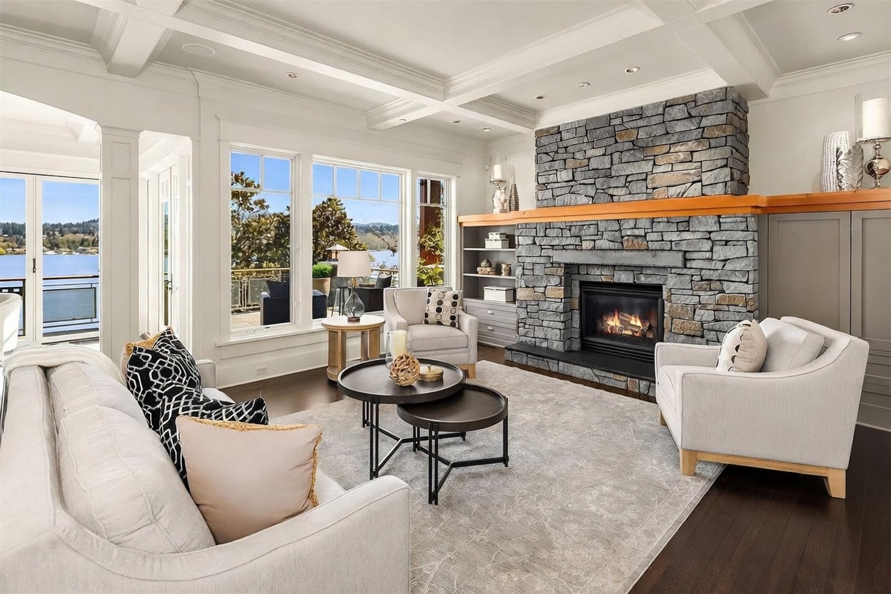This elegant living room features a grand floor-to-ceiling stone fireplace as its focal point, complemented by a coffered ceiling and rich hardwood flooring. Large windows provide an expansive, scenic view of the water and deck, creating a bright and airy atmosphere. The space is furnished with plush, neutral-toned seating and a layered rug, offering a sophisticated and inviting environment for relaxation.