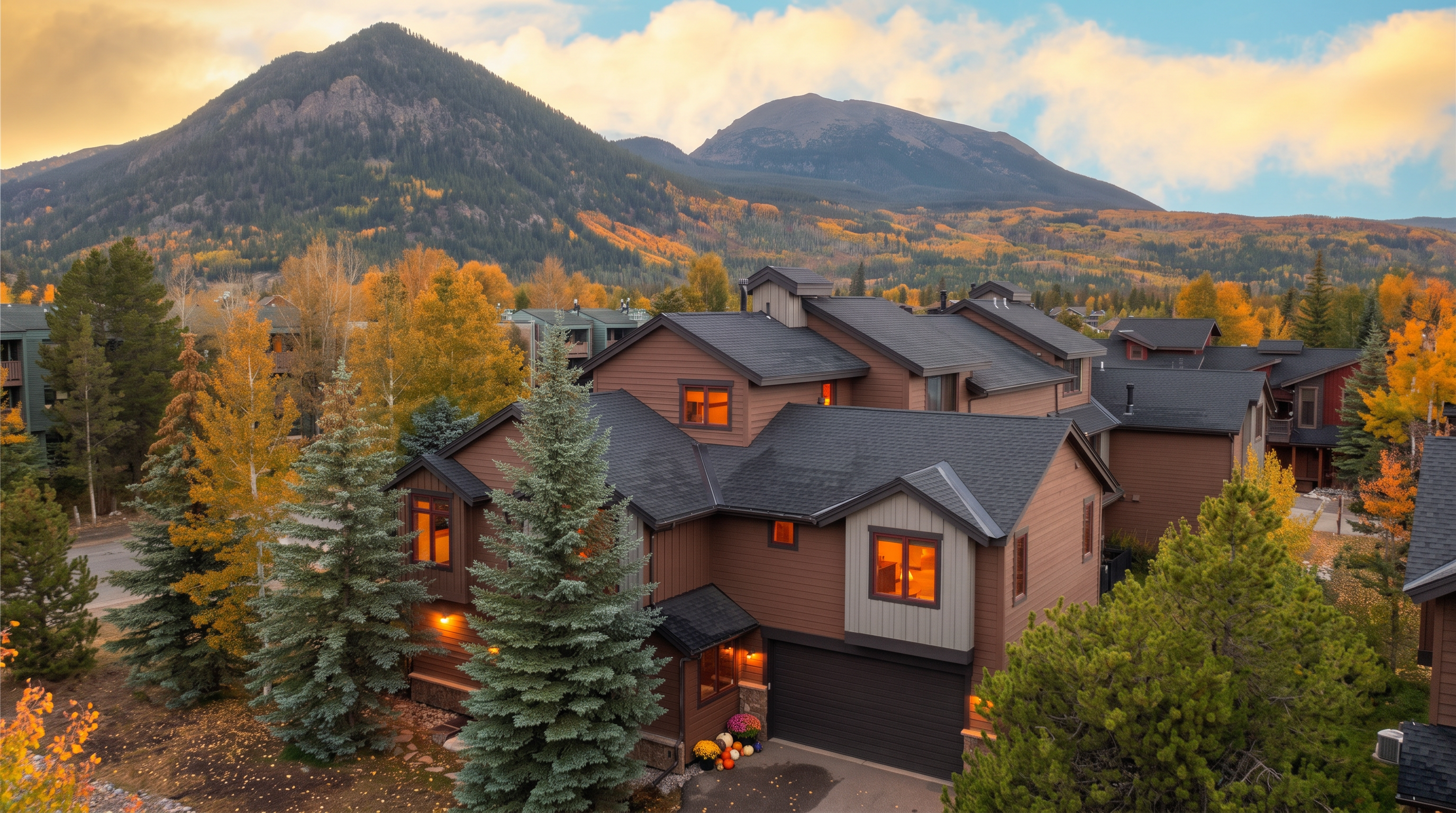 This elevated perspective captures a multi-story mountain home nestled against a backdrop of dramatic peaks and evergreen forests. The structure features a blend of brown wood siding, dark shingled roofing, and a prominent two-car garage, with warm interior lighting glowing through the windows. The setting conveys a cozy, rustic aesthetic typical of a high-altitude retreat.