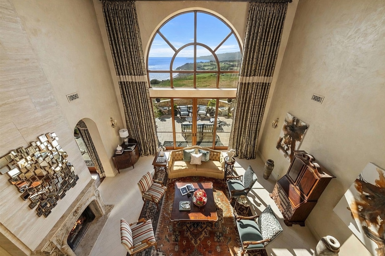 This high-angle shot captures a grand, double-height living room characterized by a massive arched window that frames a panoramic ocean view. The space is elegantly furnished with a central seating area featuring a patterned rug, a wooden coffee table, and a sofa flanked by two armchairs. To the left, a stone-clad chimney breast rises prominently, while the right side features a dark wood cabinet and a large piece of abstract wall art.