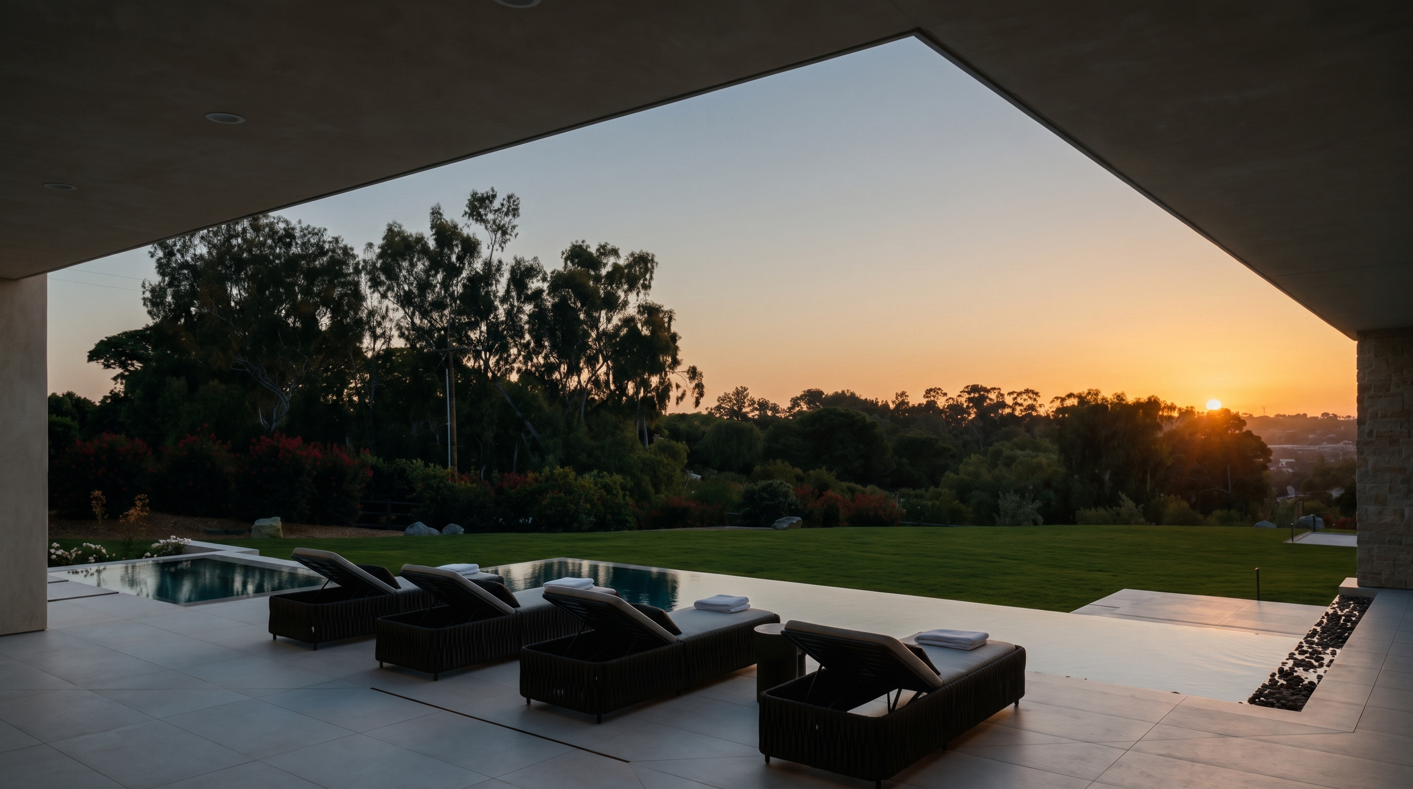 This luxurious covered patio offers a seamless transition to an expansive backyard, featuring a sleek infinity-edge pool and four modern lounge chairs arranged for sunset views. The architectural design emphasizes an open-air connection to the landscape, framed by a wide, minimalist overhang that creates a dramatic, cinematic perspective of the rolling hills and golden hour sky. The clean, neutral-toned stone flooring and refined outdoor furniture contribute to a sophisticated, resort-like atmosphere.