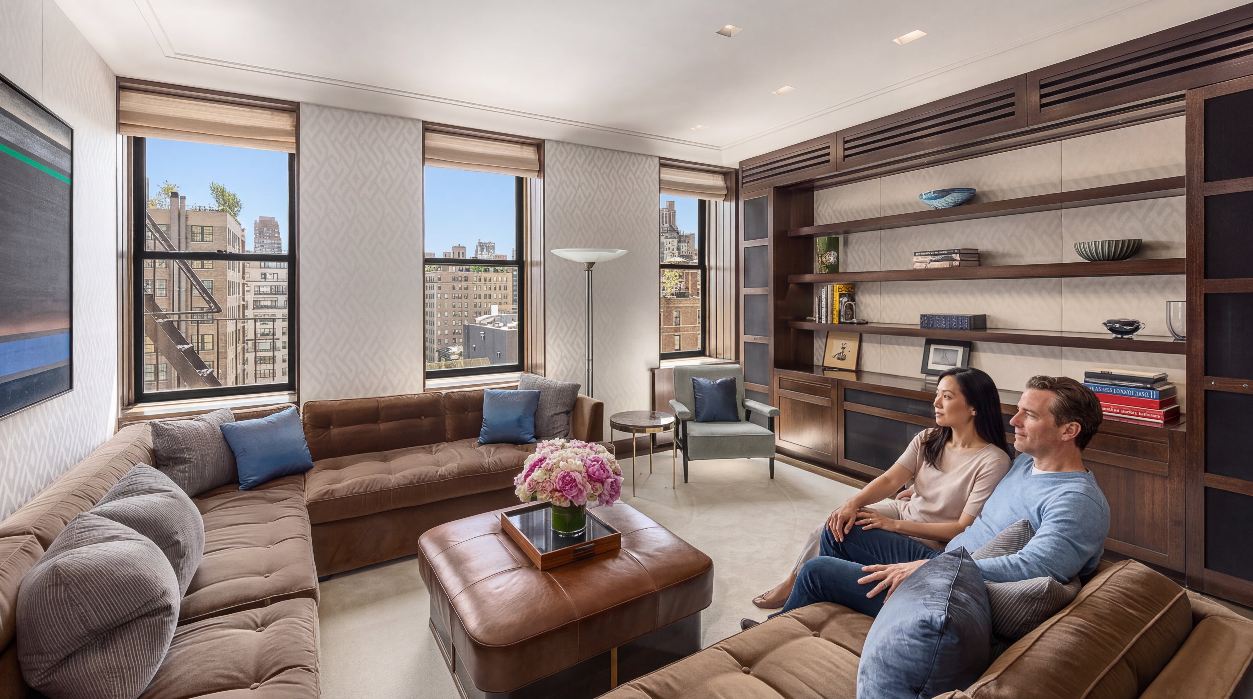 This sophisticated living room features a large, plush brown leather sectional sofa, a matching ottoman, and expansive windows that offer clear, urban skyline views. The space is anchored by a custom floor-to-ceiling wooden built-in shelving unit and finished with neutral, textured wallpaper and contemporary accents, creating an elegant and comfortable atmosphere.