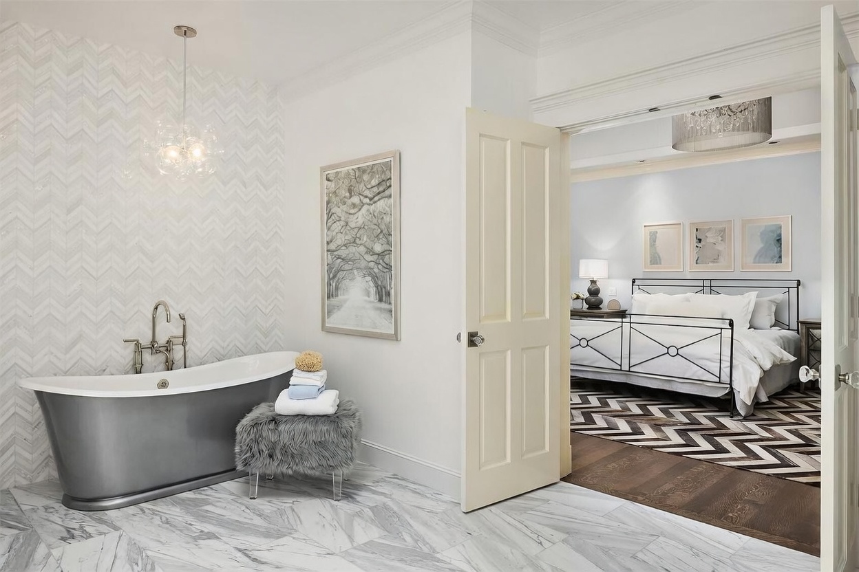 This elegant primary bathroom features a freestanding soaking tub set against a wall with a decorative herringbone-patterned tile backsplash. A plush, grey faux-fur stool sits beside the tub, while a doorway provides a direct view into an adjacent bedroom with a metal-framed bed and patterned area rug. The space is finished with light-toned stone flooring and a modern glass pendant light, creating a sophisticated and serene atmosphere.