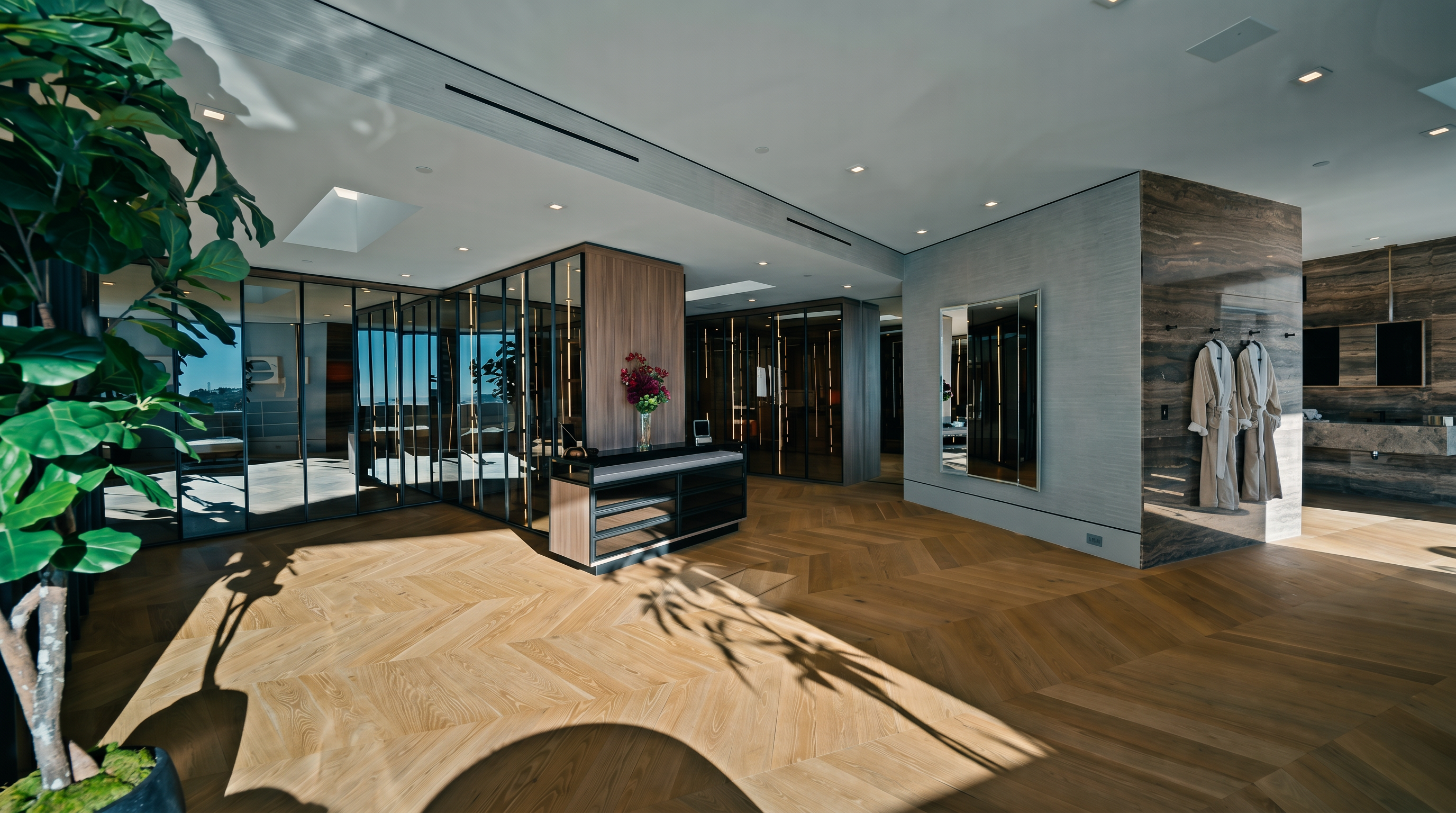 This expansive, high-end walk-in closet features light-toned herringbone wood flooring and a sophisticated layout with glass-enclosed storage units. The space is anchored by a central island with a dark countertop and floral accents, while a marble-clad wall displays hanging robes, suggesting a luxurious, boutique-like atmosphere. The perspective is wide and inviting, showcasing the seamless integration of modern design and functional storage.