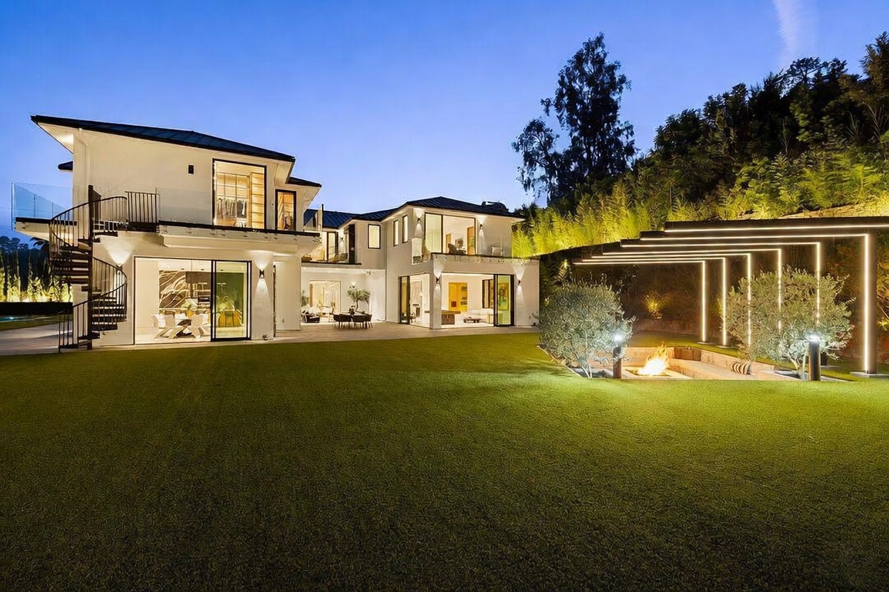 This stunning contemporary residence is captured from the rear yard at twilight, showcasing a multi-level architectural design with expansive glass walls and a spiral staircase. The manicured lawn leads to a sunken fire pit area framed by a modern, illuminated pergola structure. The warm glow from the interior lighting contrasts beautifully with the cool evening sky, highlighting the home's sophisticated indoor-outdoor living capabilities.