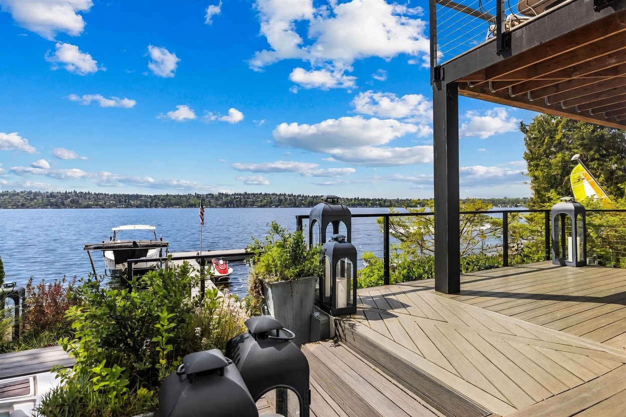 This elevated wooden deck offers a serene, panoramic view of a large lake under a bright, partly cloudy sky. The space is furnished with decorative black lanterns and potted greenery, creating a sophisticated outdoor living area that overlooks a private dock with a boat and personal watercraft. The perspective captures the seamless transition between the home's architectural structure and the expansive waterfront scenery.