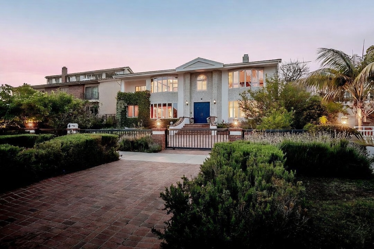 This two-story residence features a symmetrical facade with a light-colored brick exterior and a prominent central entryway with a blue double door. The property is framed by lush, mature landscaping, including manicured hedges and a palm tree, and is secured by a black metal gate. A brick-paved driveway leads up to the entrance, creating a welcoming and established curb appeal during the soft light of dusk.