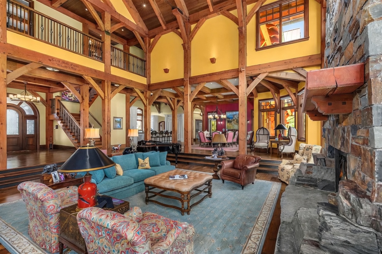 This expansive, open-concept living area features a grand timber-frame structure with exposed beams and high vaulted ceilings. The room is anchored by a massive stone-clad chimney breast and includes a comfortable seating area with a teal sofa, patterned armchairs, and a large leather ottoman. Beyond the living space, a raised dining area and a glimpse of a kitchen are visible, all unified by warm wood flooring and a bright, inviting atmosphere.