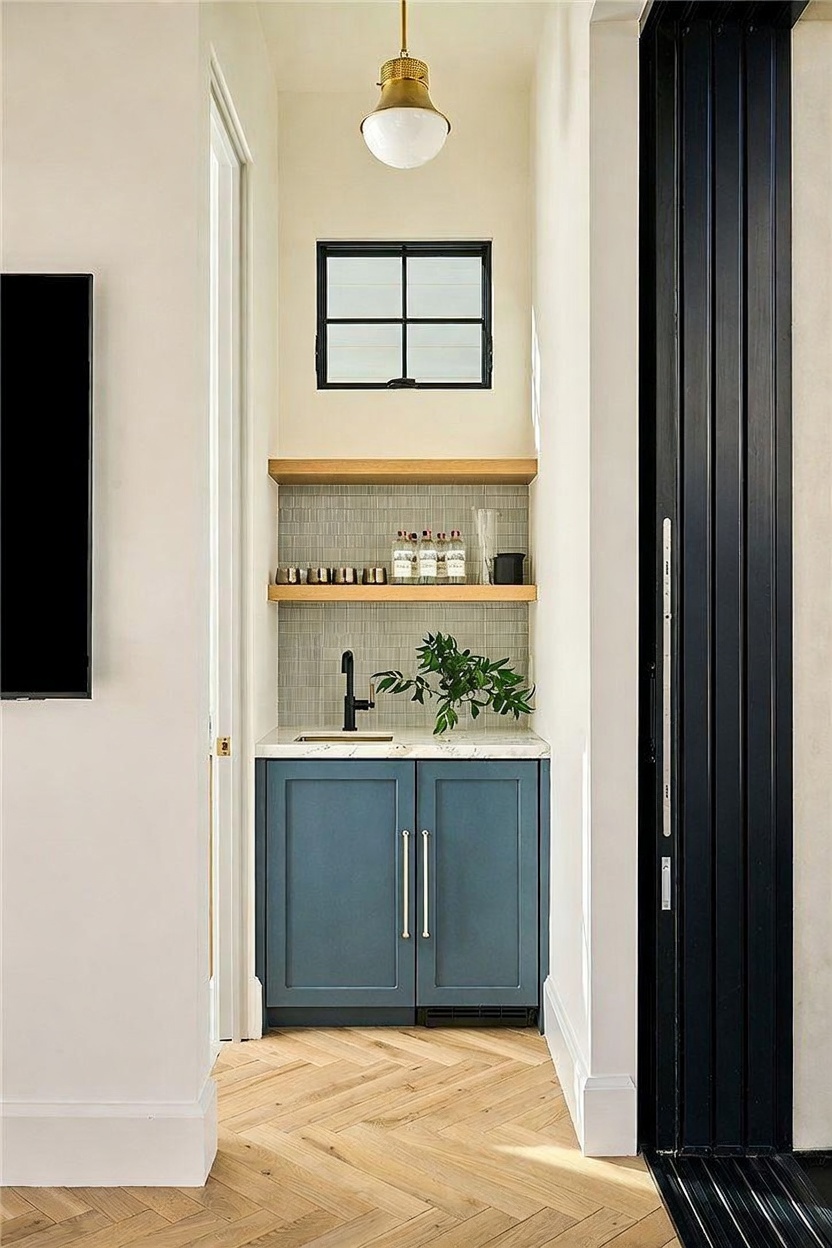 This image showcases a sophisticated wet bar nook featuring deep blue cabinetry topped with a light-toned stone counter and a sleek black faucet. Two light wood floating shelves are mounted against a textured tile backsplash, providing an elegant display area beneath a small, grid-paned window. The space is finished with light-toned herringbone wood flooring and illuminated by a single brass pendant light, creating a refined and functional transition area.