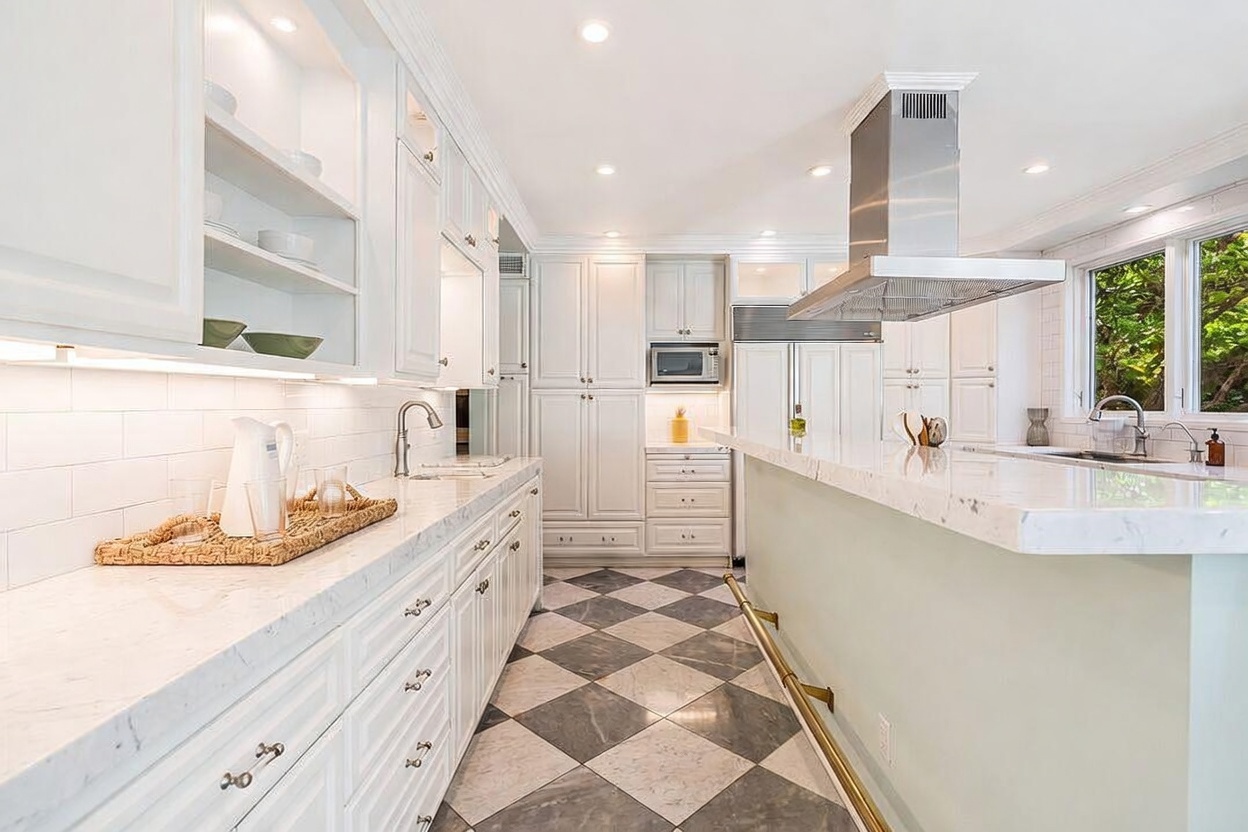 This bright and elegant kitchen features white cabinetry, stone countertops, and a central island with a breakfast bar and brass foot rail. The space is anchored by a striking black and white checkered tile floor and a stainless steel range hood above the island. Large windows allow natural light to fill the room, creating an airy and sophisticated atmosphere.