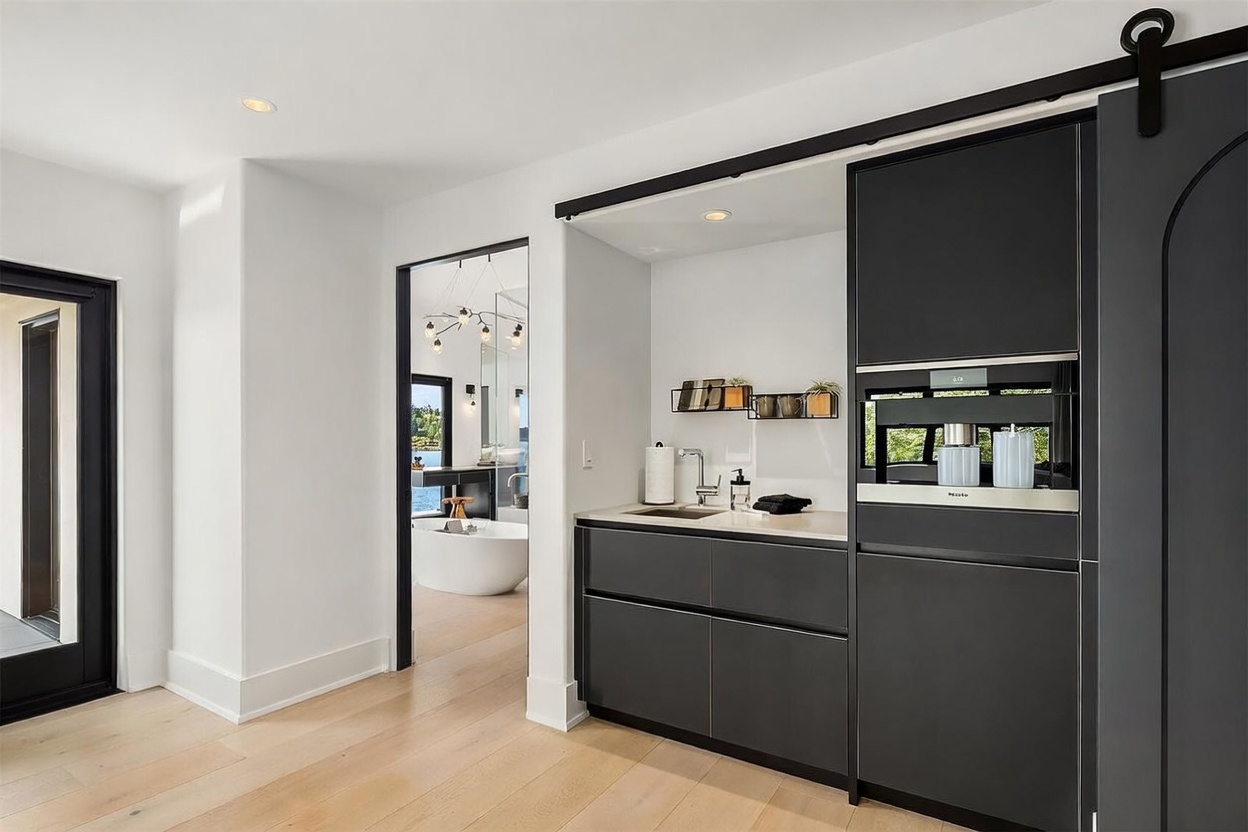 This modern kitchenette area features sleek, dark cabinetry and a built-in Miele coffee system, set against a crisp white wall. A sliding barn door track is visible above the unit, while a doorway leads into a bright, adjacent bathroom featuring a freestanding tub. The space is finished with light wood flooring, creating a sophisticated and minimalist aesthetic.