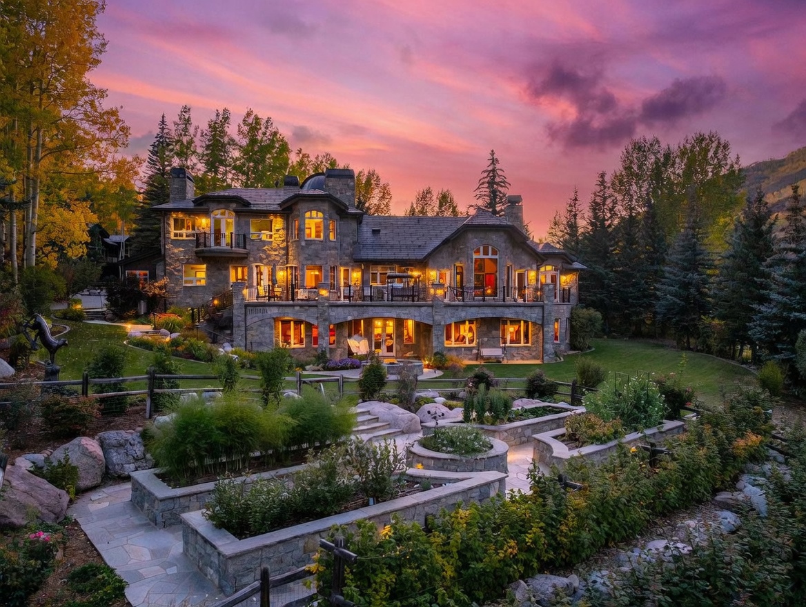 This stunning multi-story stone estate is captured at dusk, showcasing a vibrant sunset sky behind the property. The rear facade features a spacious elevated deck with iron railings, a lower-level covered patio, and a meticulously landscaped terraced garden with stone retaining walls. The warm glow from the interior lighting creates an inviting atmosphere, highlighting the architectural detail and the expansive outdoor living spaces.