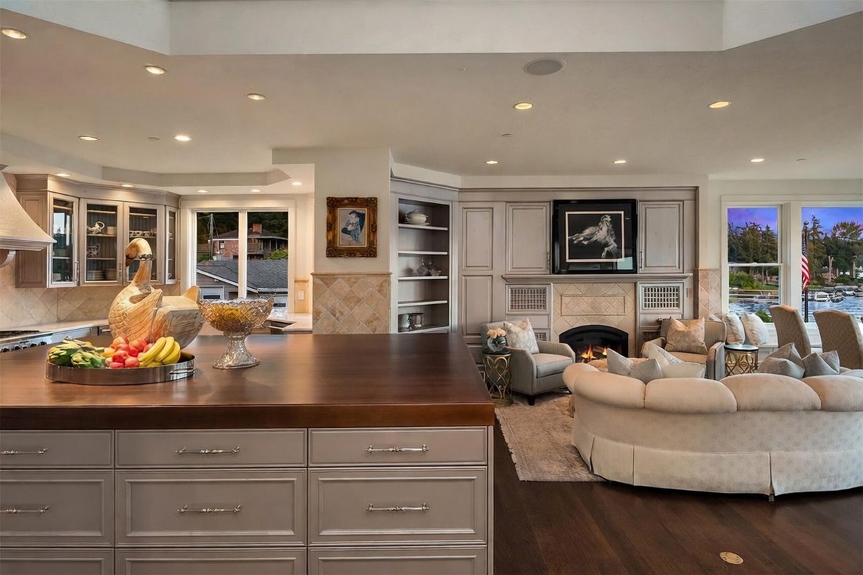 This open-concept space seamlessly blends a kitchen with a cozy living area, featuring a large island with a dark wood countertop and light-toned cabinetry. A fireplace with a decorative surround serves as a focal point in the living area, which is furnished with a curved sofa and armchairs. Large windows provide ample natural light and views of the outdoors, creating an inviting and sophisticated atmosphere.