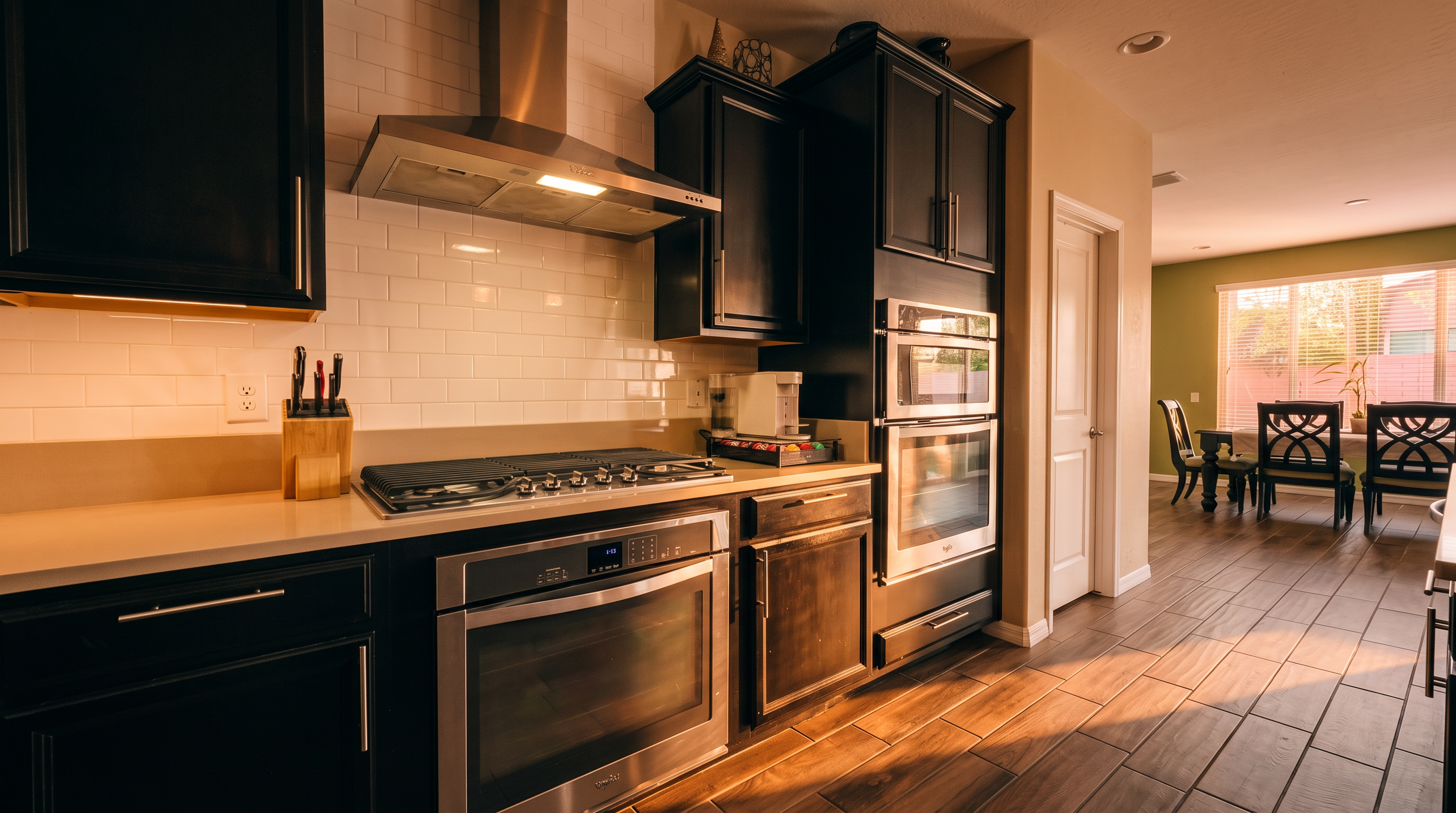 This modern kitchen features dark cabinetry paired with light-toned countertops and a white subway tile backsplash. The space is equipped with stainless steel appliances, including a gas cooktop, a built-in oven, and a microwave, all set against a neutral color palette. The perspective captures the functional workspace, with a view leading into an adjacent dining area featuring dark wood-toned flooring.